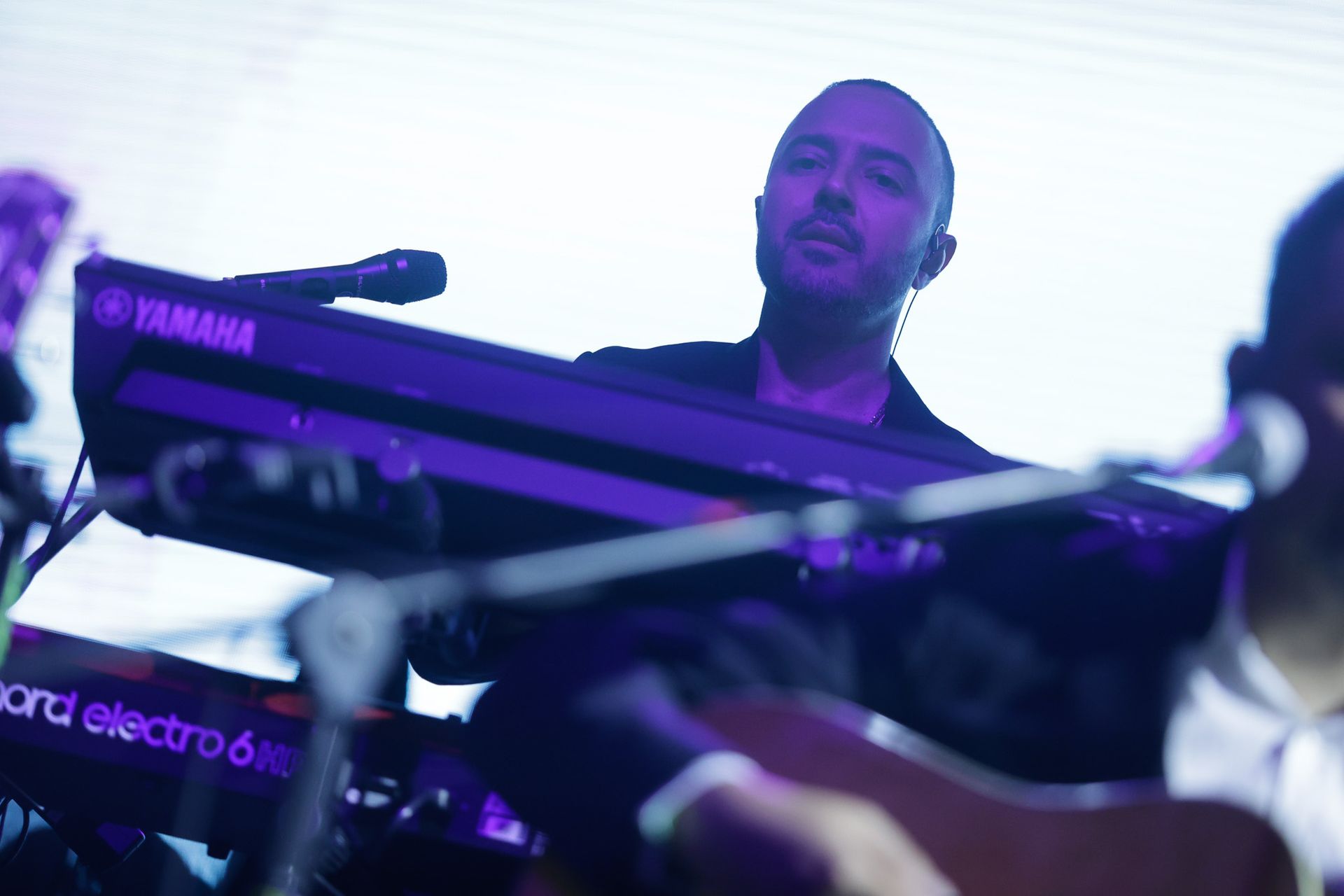 Tecladista se apresentando ao vivo no palco, iluminado com tons roxos. Um microfone está posicionado próximo ao seu rosto.