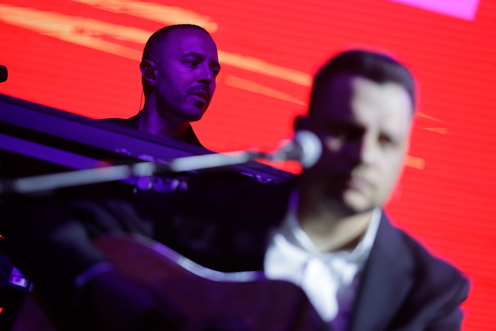 Dois homens se apresentando no palco. Um toca teclado, o outro canta ao microfone. Fundo vermelho.