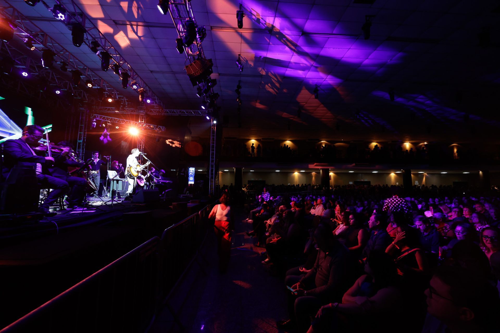 Banda se apresentando no palco, banhada por luzes de palco roxas e laranjas, com uma plateia sentada à frente.
