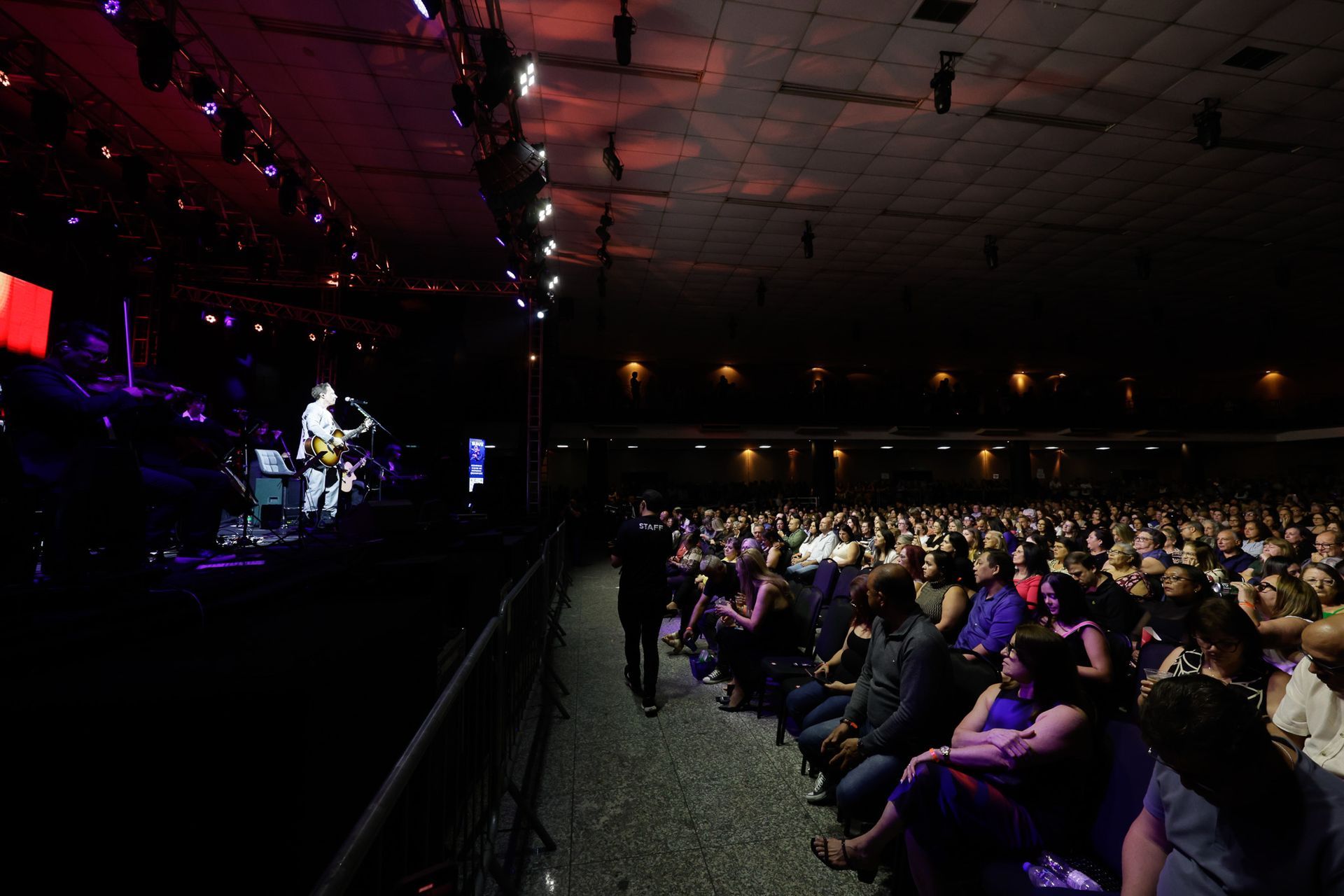 Cena de concerto: Banda se apresentando no palco, grande público assistindo. Local escuro com luzes de palco.