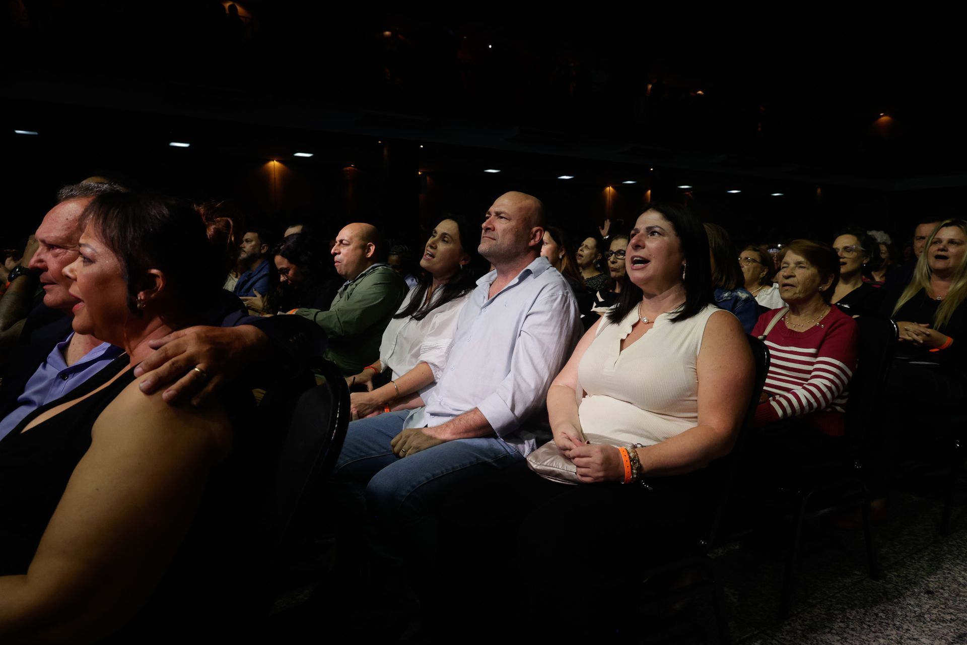 Público em teatro escuro, assistindo e reagindo. Alguns sorriem, outros cantam junto.