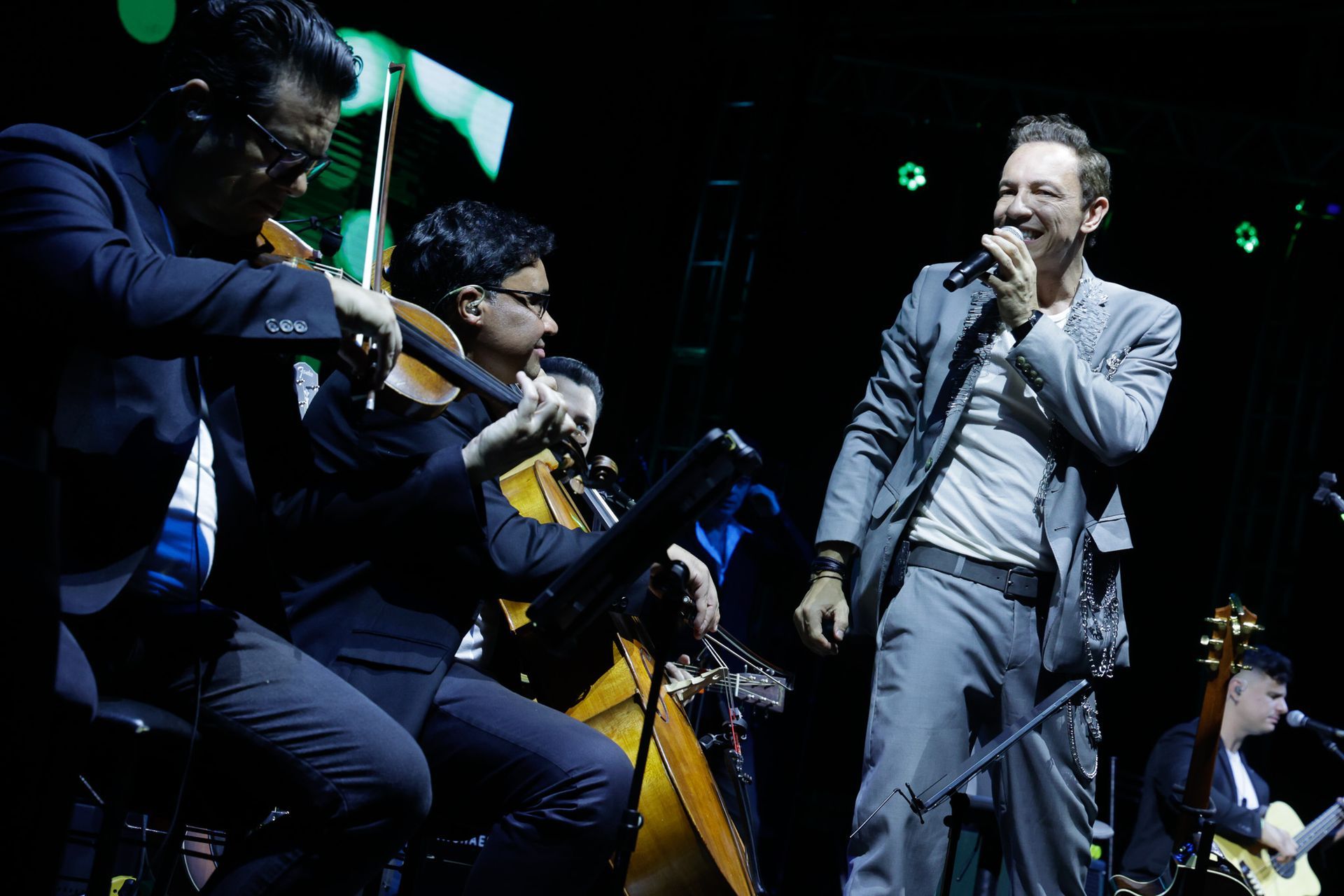 Um artista cantando ao microfone no palco com músicos de cordas em um ambiente com pouca luz.