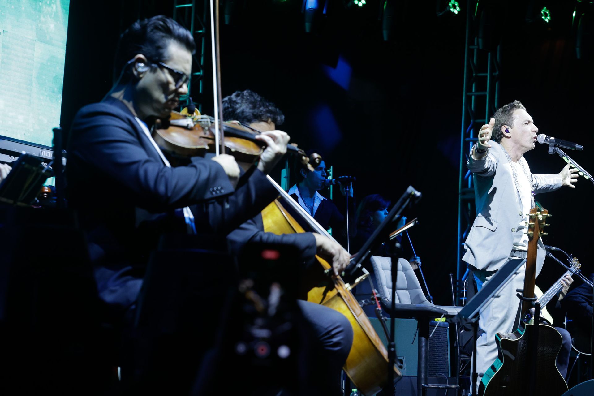 Músico no palco com violinistas e violoncelo; cantor de terno se apresenta com os braços estendidos.