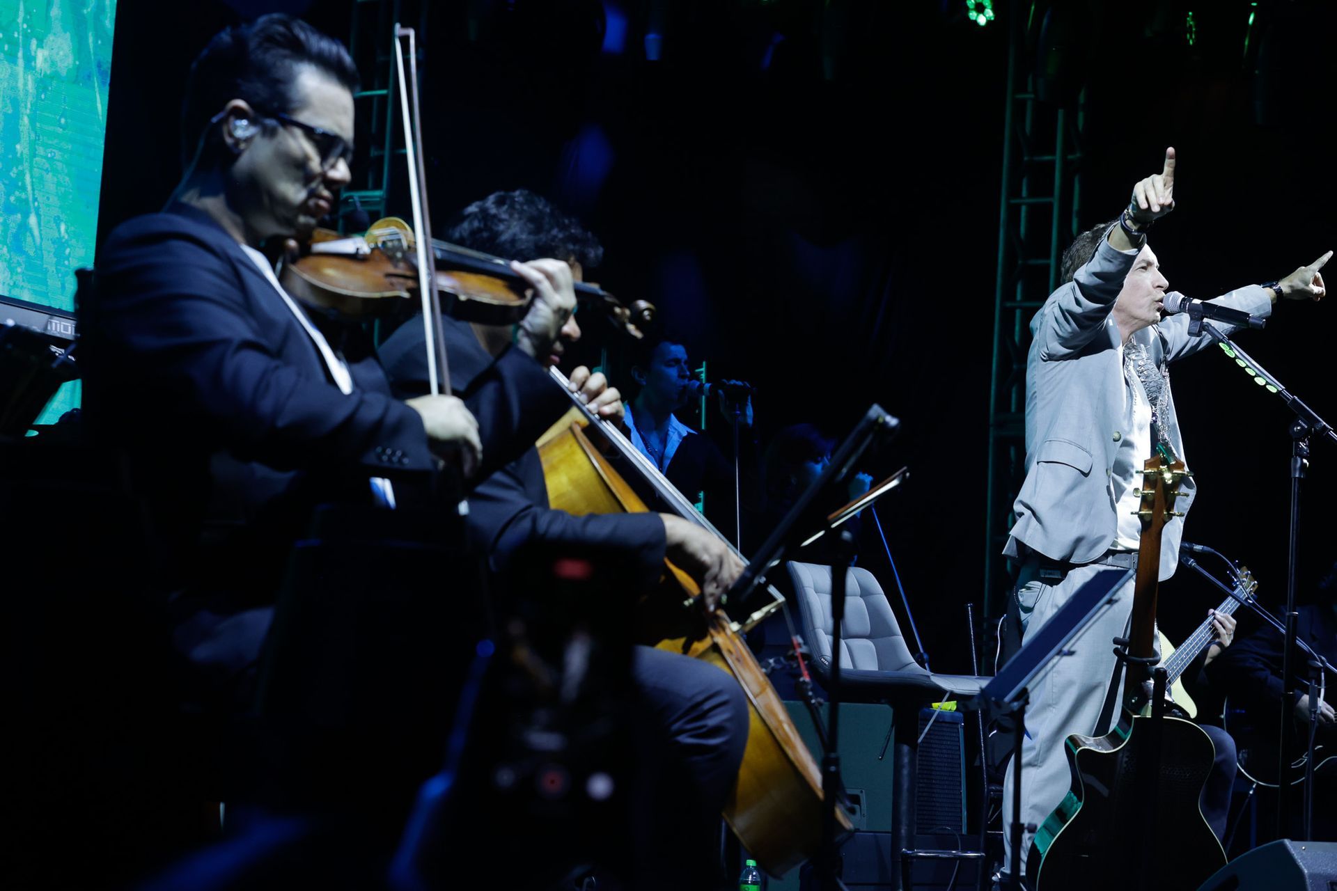 Músicos se apresentando no palco: violinista, violoncelista e cantor com microfone. O palco está escuro.