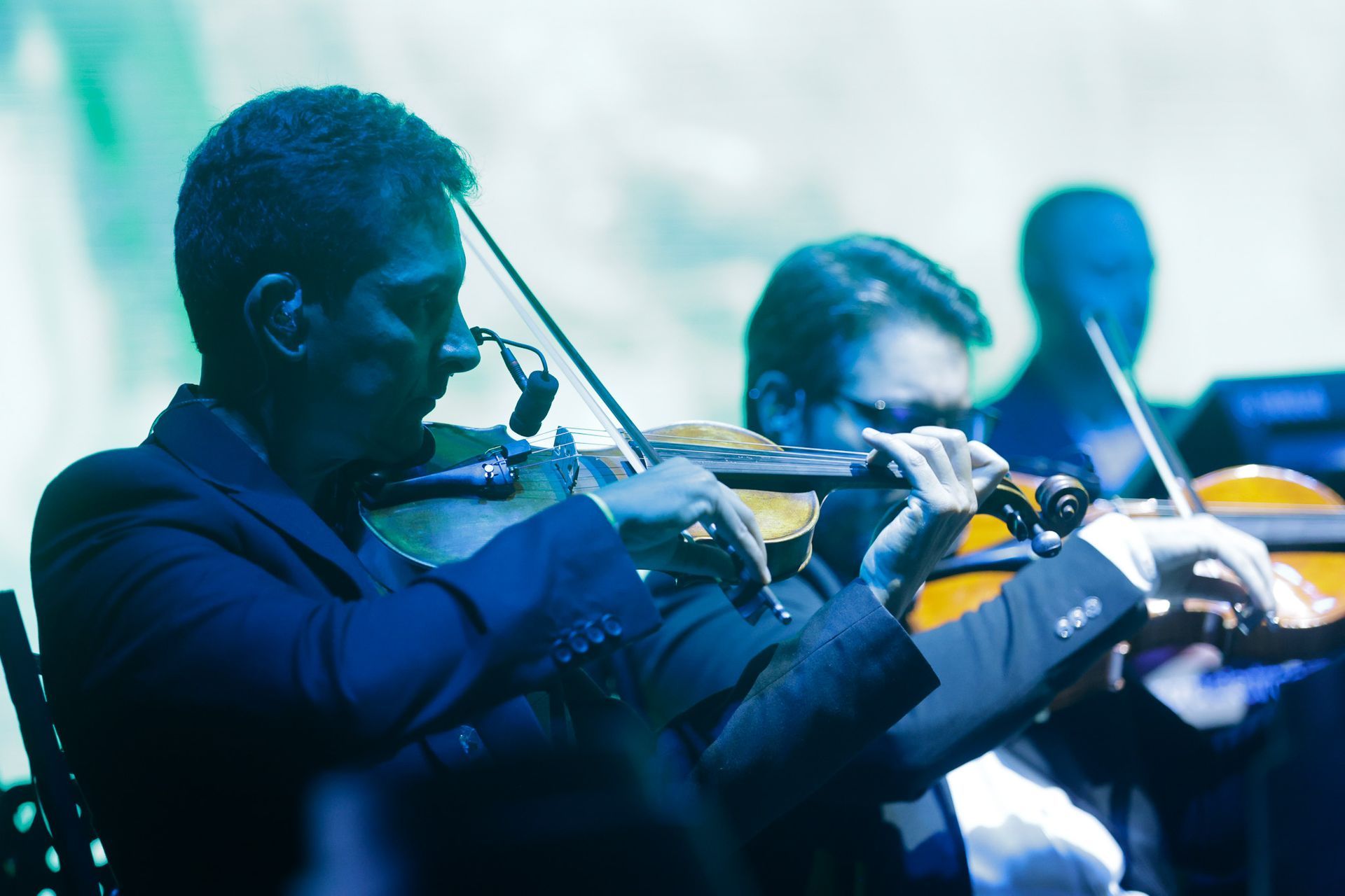 Músicos tocando violinos em uma orquestra, palco iluminado com tons de azul e verde.