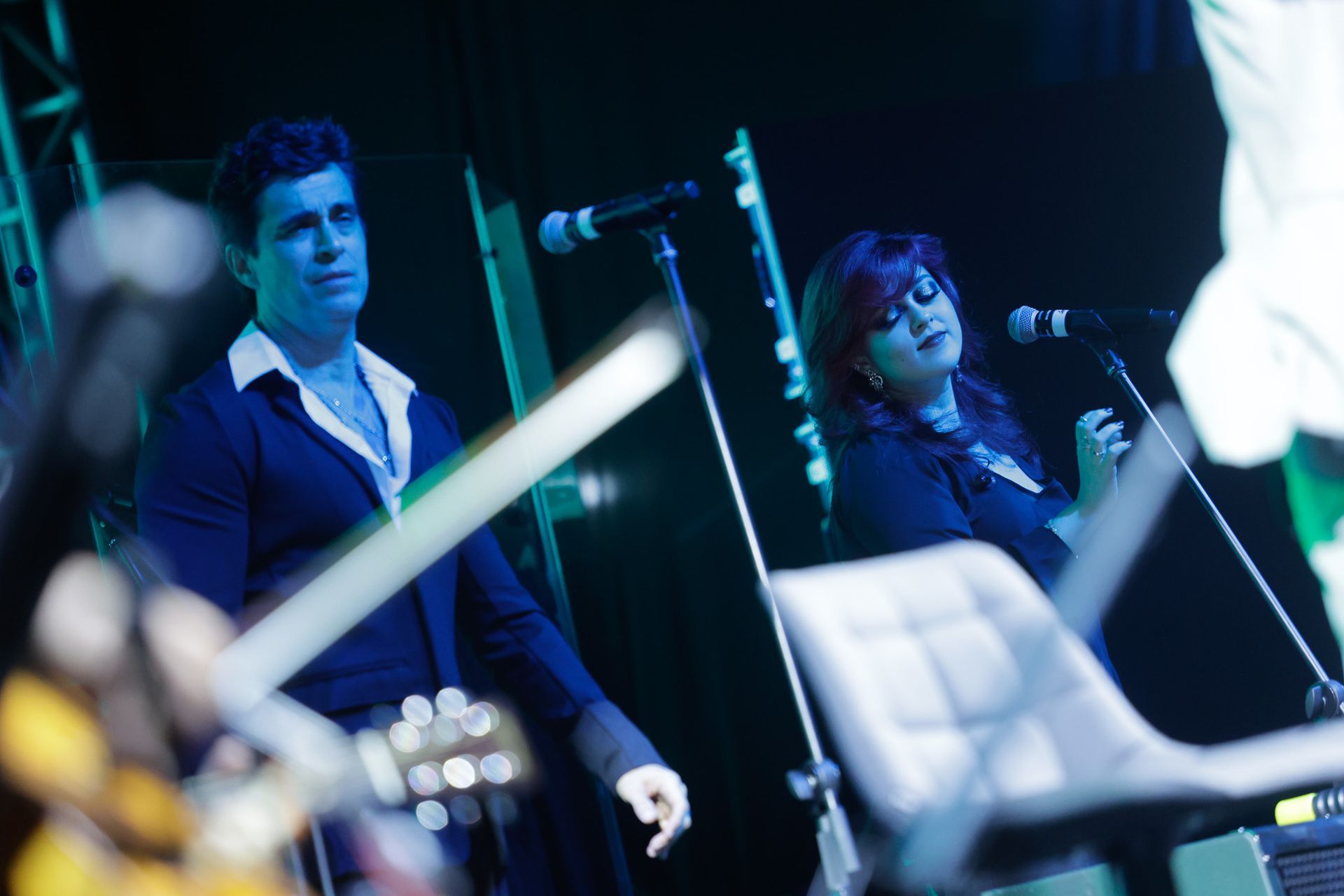Dois cantores no palco, um homem e uma mulher, apresentando-se com microfones sob iluminação cênica azul.