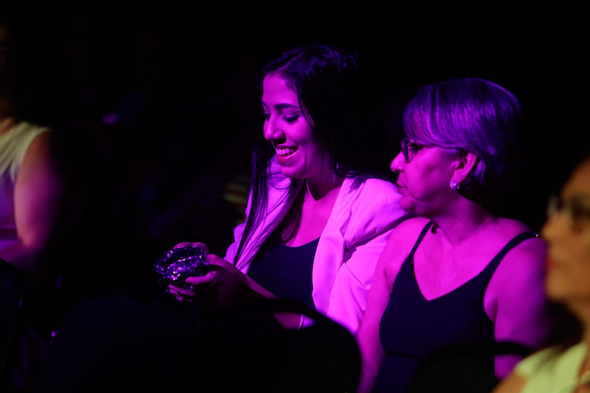 Duas mulheres sorrindo, iluminadas por luz roxa. Uma segura um pequeno objeto, a outra observa. Ambiente interno.