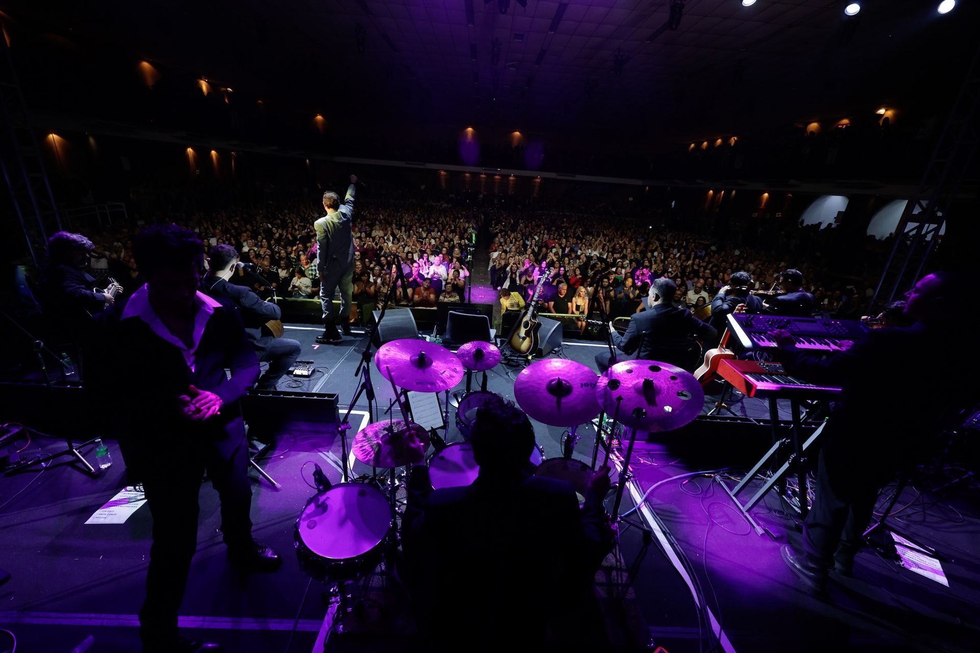 Banda se apresentando no palco diante de uma grande plateia, banhada por luzes roxas.