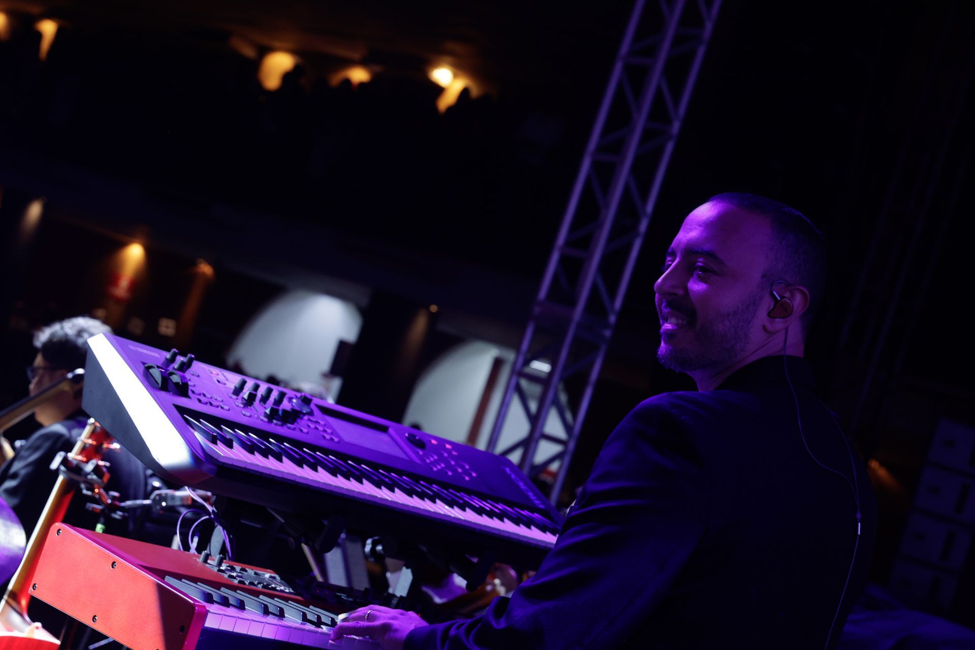 Tecladista sorridente, tocando no palco com teclados roxos e vermelhos, em ambiente escuro.