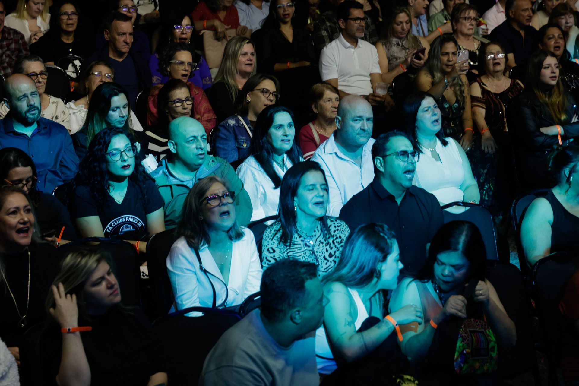 Público assistindo a uma apresentação, muitos com a boca aberta. Iluminação cênica azul, fundo escuro.