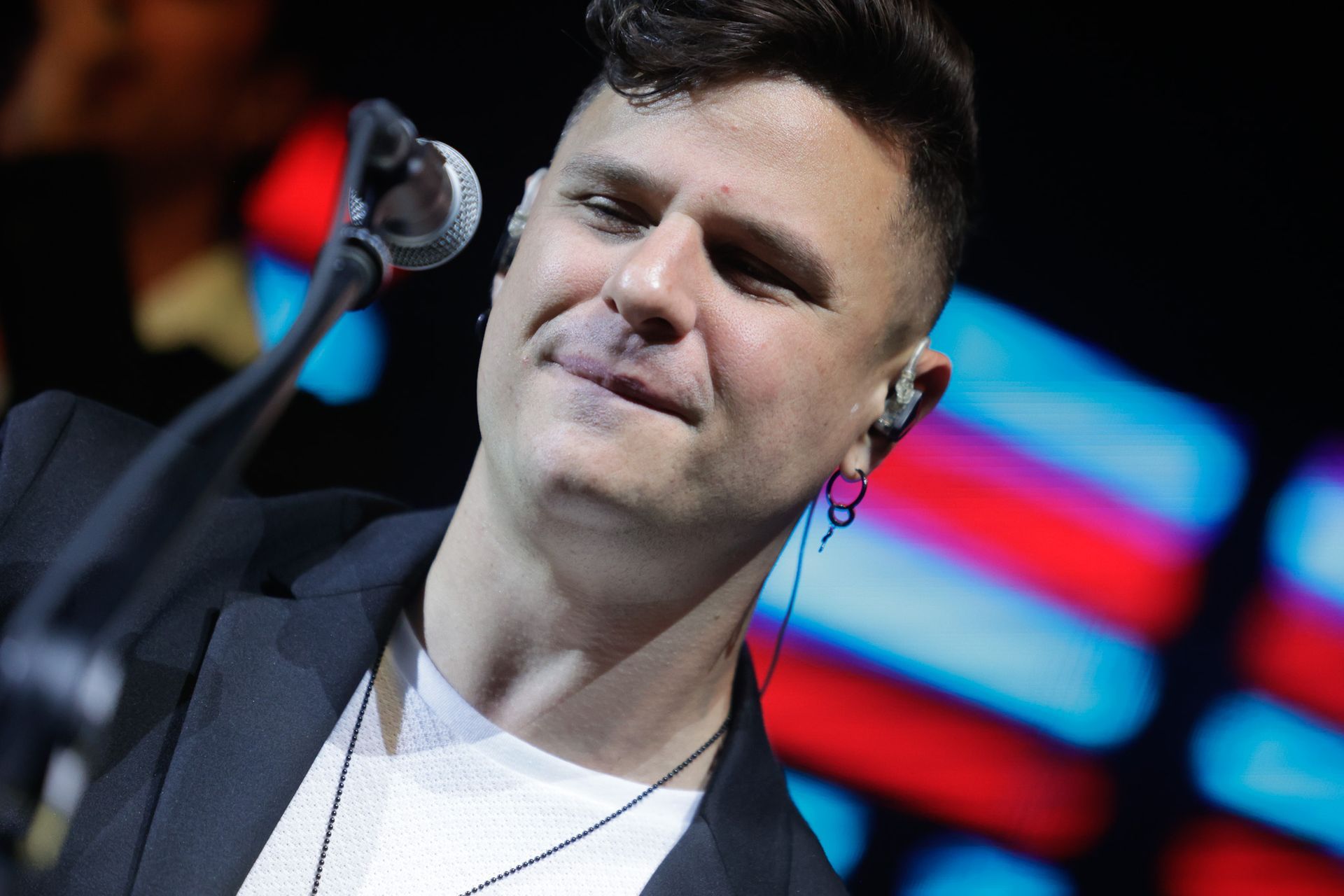 Um artista de cabelo curto e escuro, vestindo terno, cantando ao microfone no palco, sorrindo.