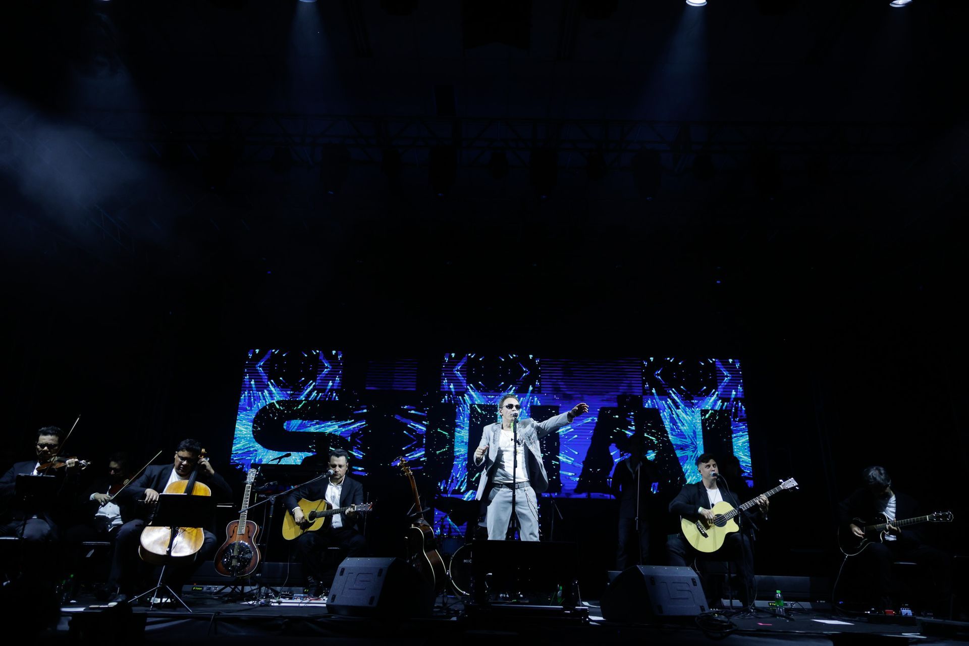 Banda se apresentando no palco com um grande painel de fundo com as iniciais 
