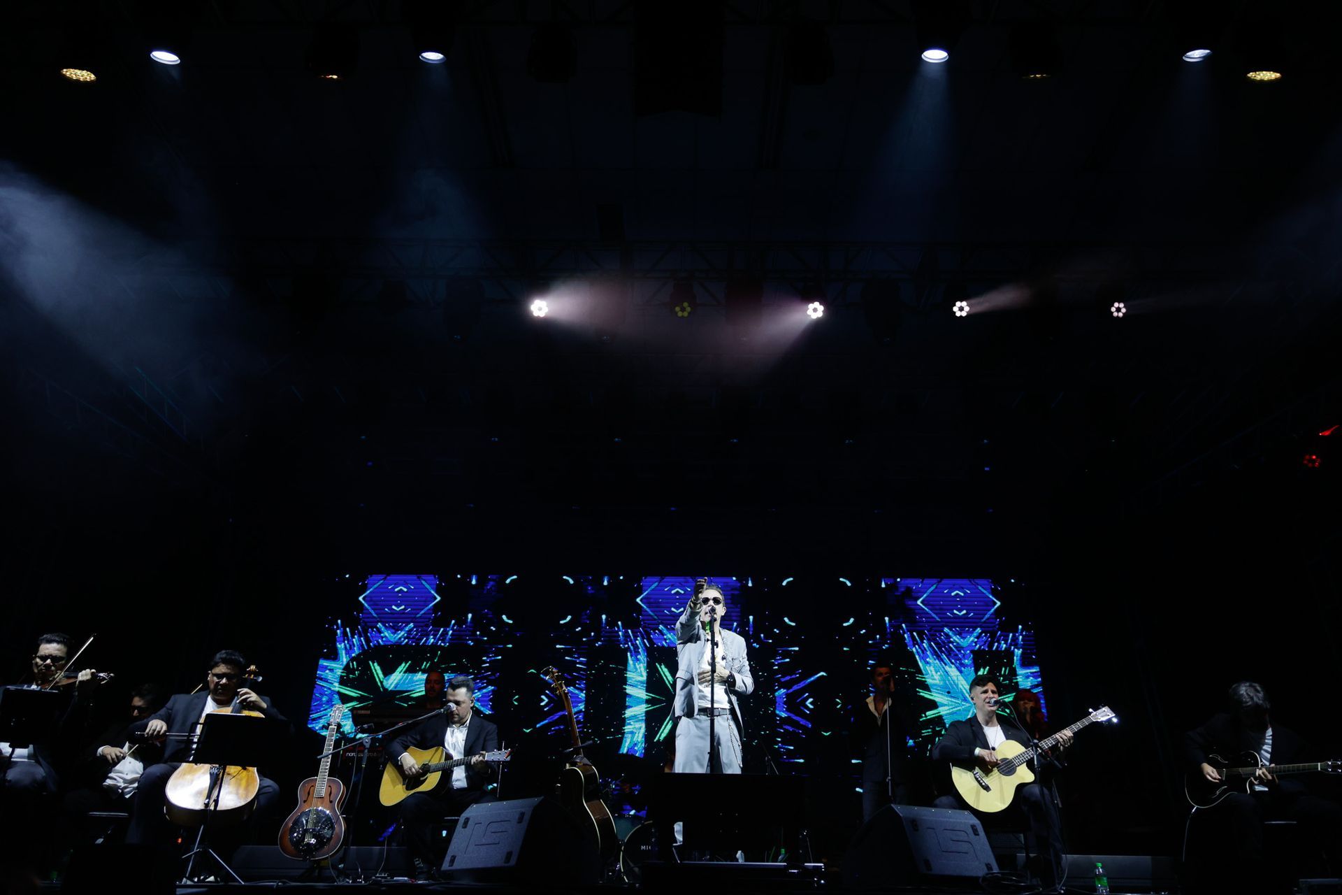Um artista de terno branco canta no palco com uma banda e um telão em um local escuro.