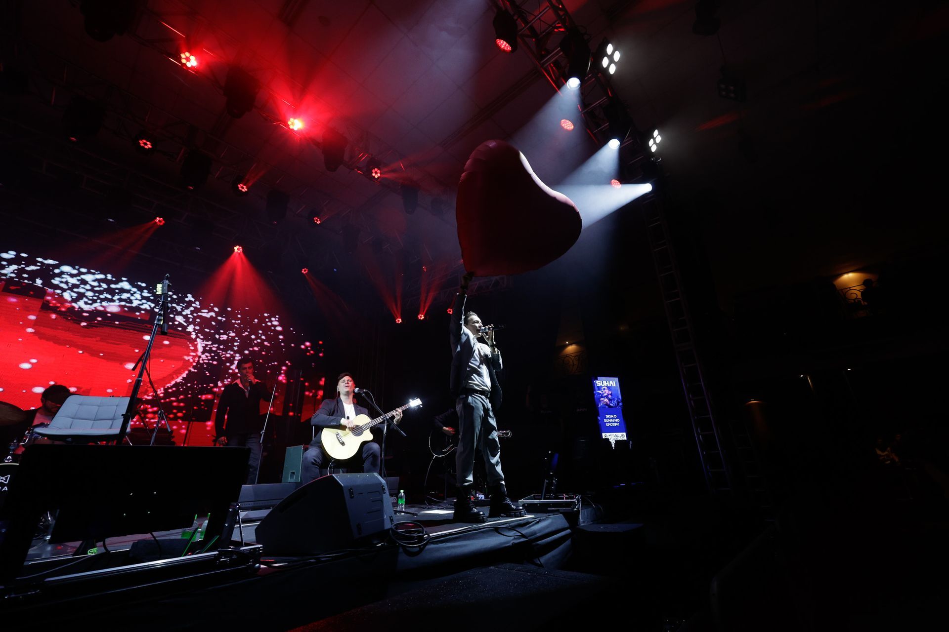 Cantor no palco, segurando um balão em forma de coração, se apresentando com a banda, iluminação vermelha no palco.