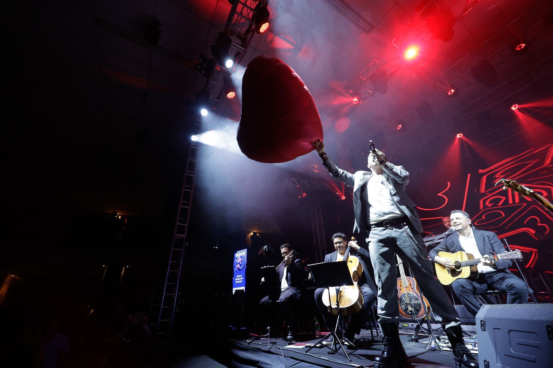 Um músico no palco com um balão em forma de coração, a banda atrás, luzes vermelhas no palco.