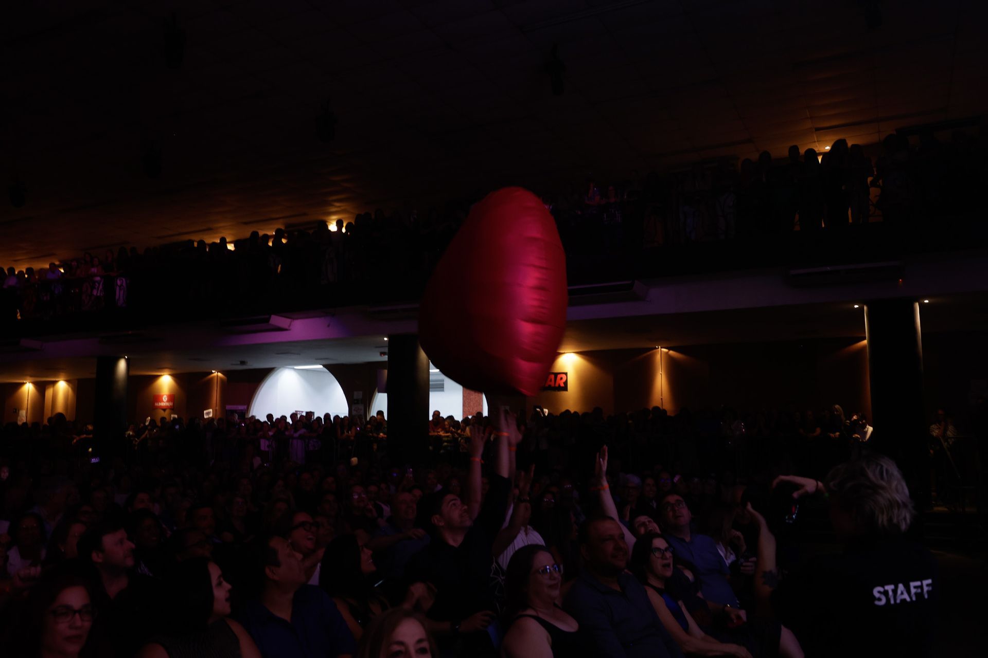 Público em um show; uma pessoa segura um grande balão vermelho em forma de coração. Local escuro com iluminação ambiente.
