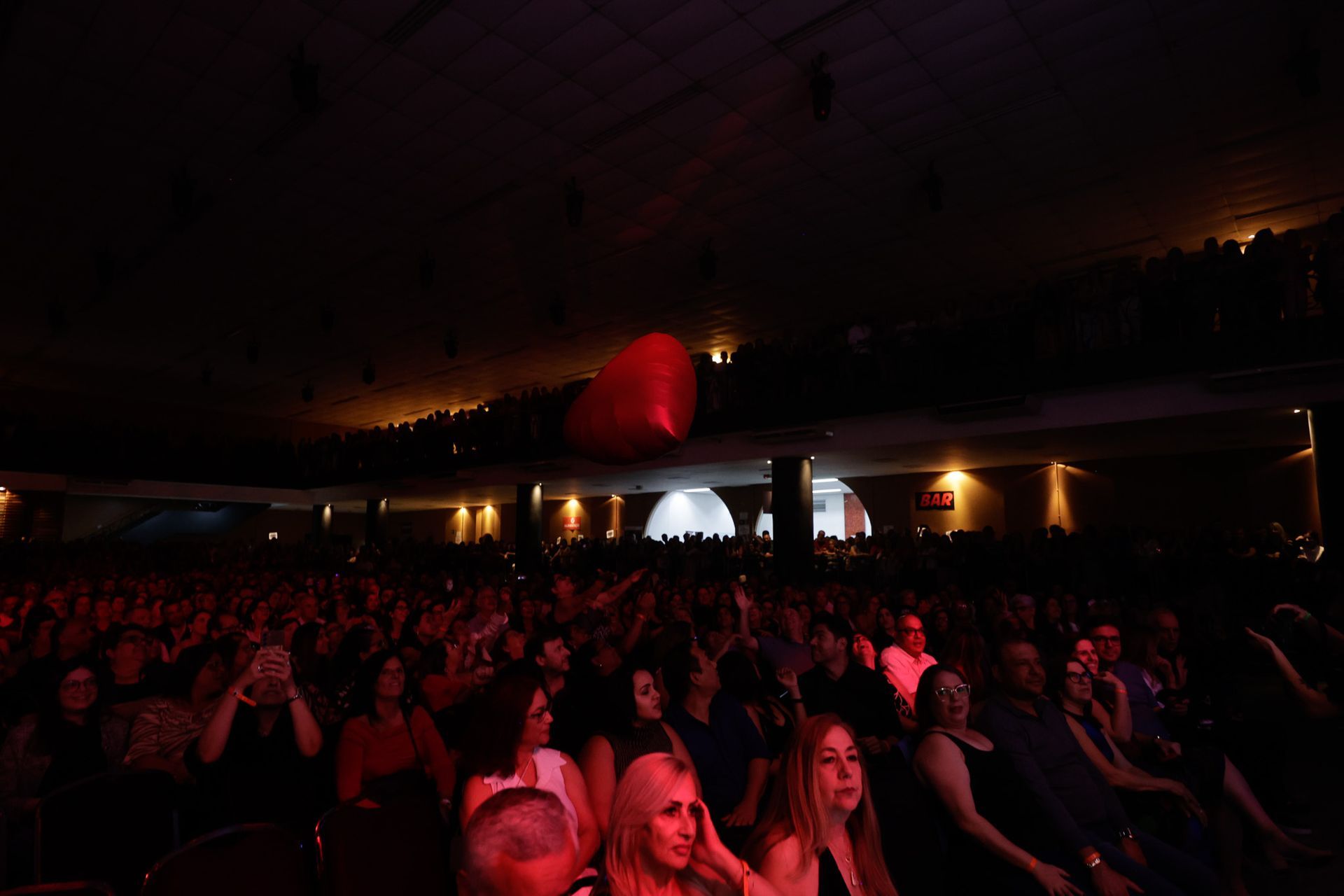 O público assiste a uma apresentação em um teatro com pouca luz. Um balão vermelho em forma de coração flutua acima.