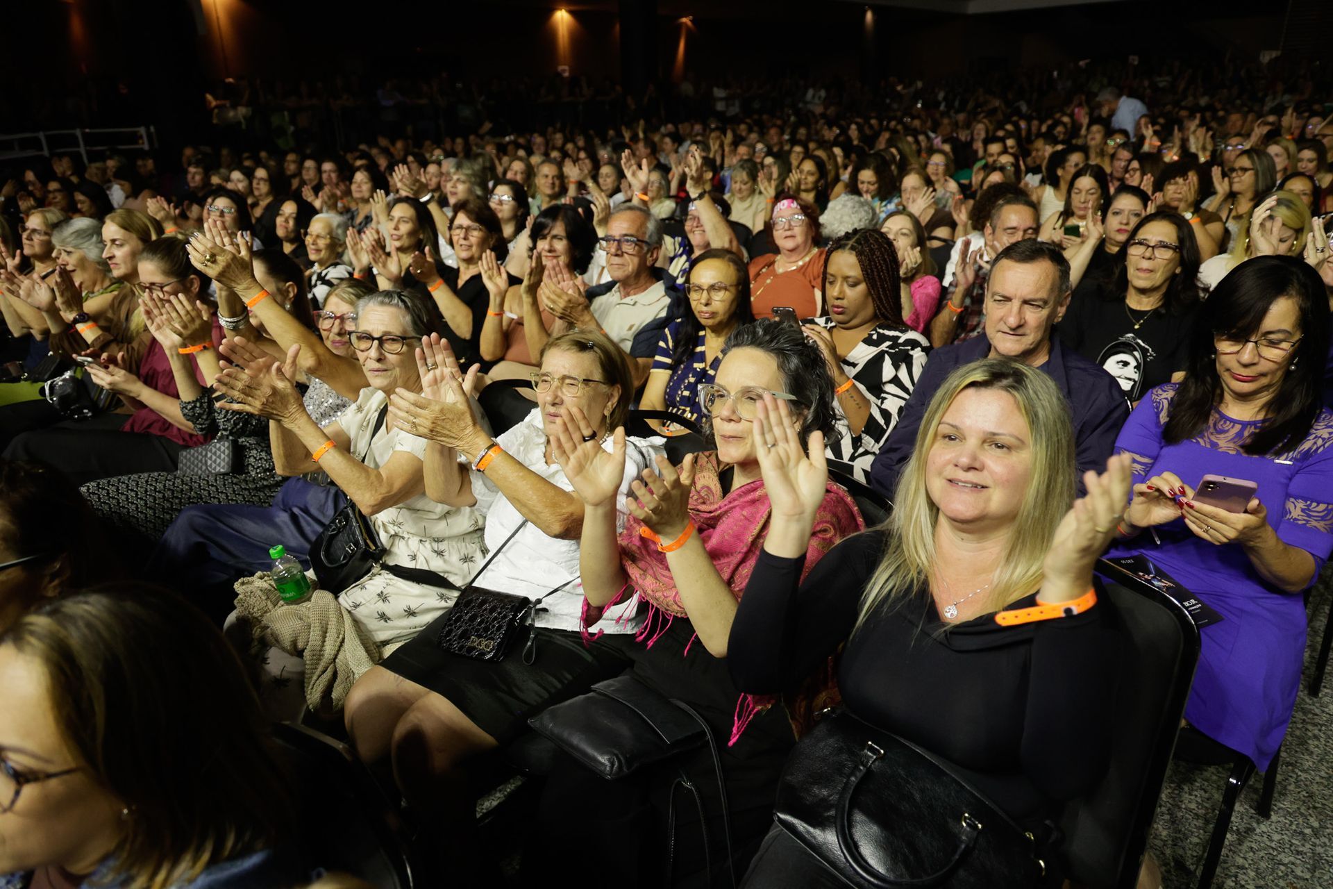 Em um auditório com iluminação tênue, a plateia aplaude com as mãos erguidas.