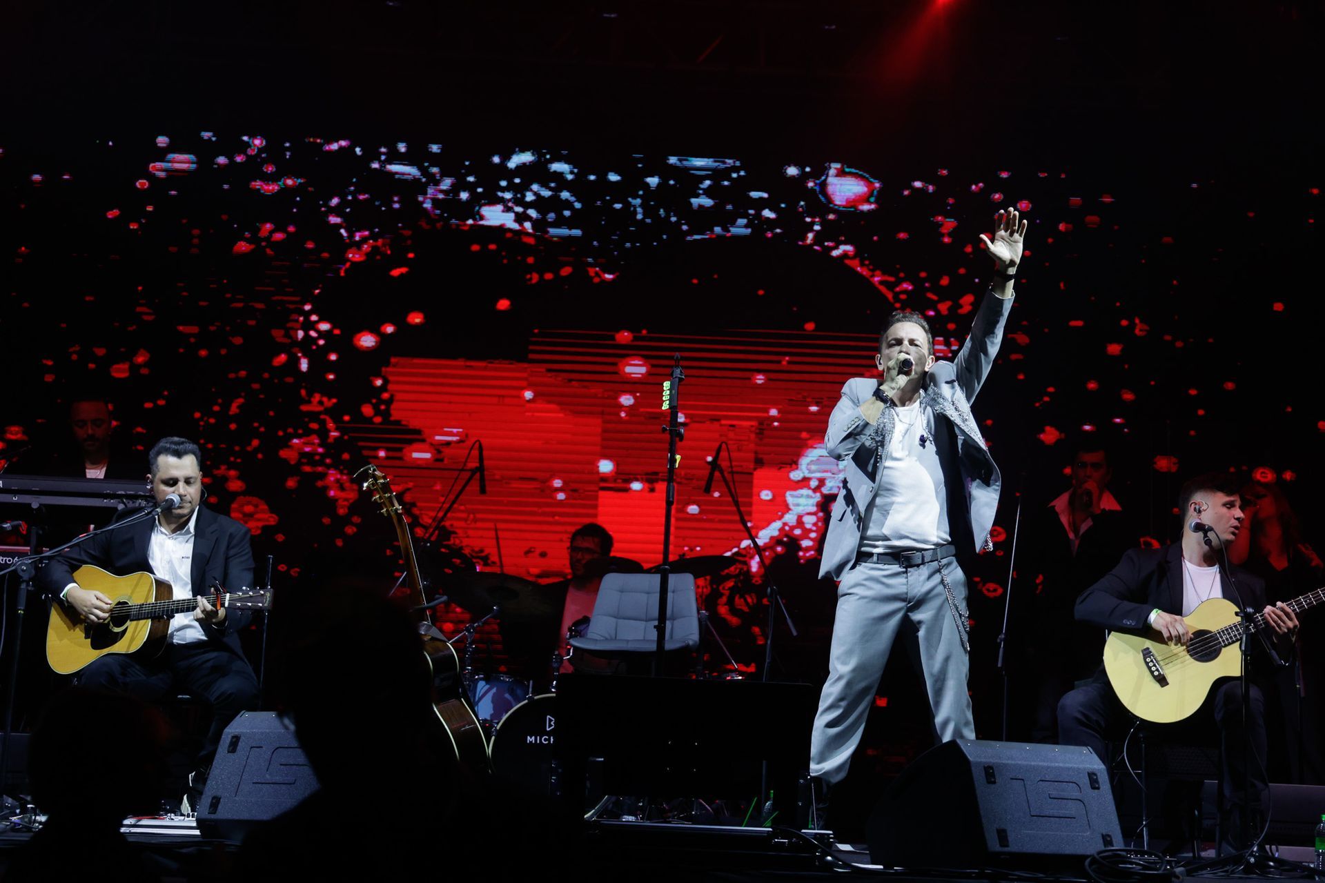 Um cantor com os braços erguidos se apresenta no palco acompanhado por dois guitarristas. Fundo vermelho com músicos.