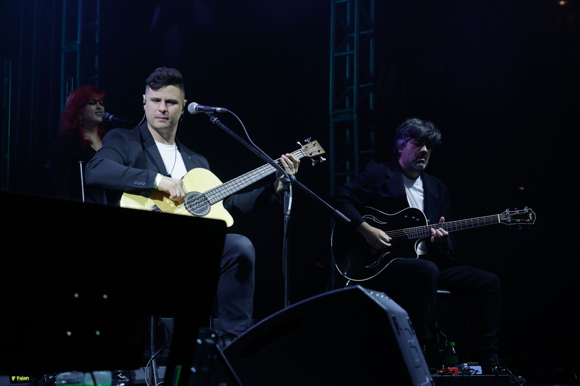 Dois homens tocando guitarra no palco, se apresentando em um local escuro.