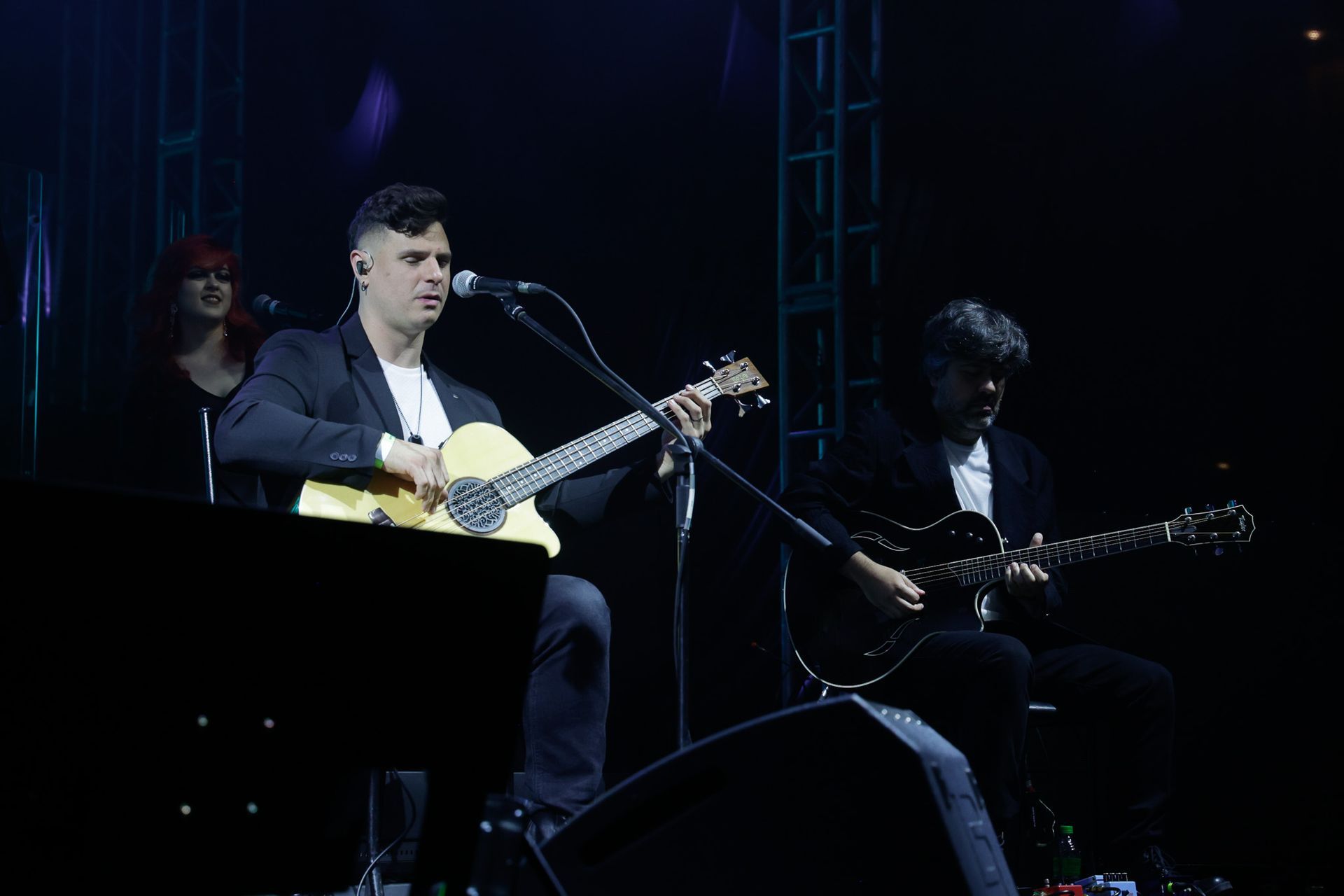 Uma banda se apresenta no palco. Um guitarrista canta ao microfone, enquanto outro guitarrista toca