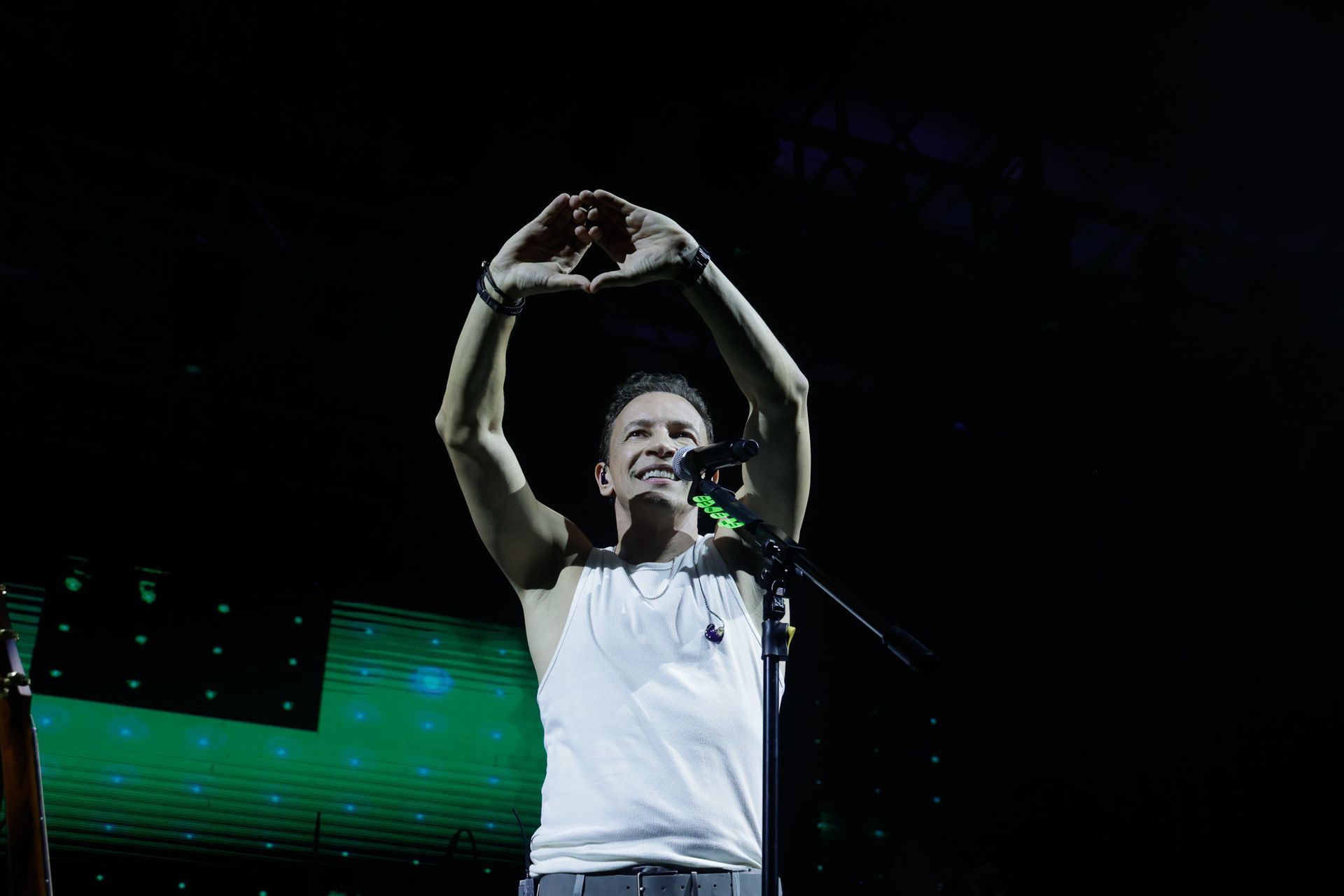 Um artista no palco com os braços erguidos, as mãos formando um coração. Regata branca, fundo escuro.
