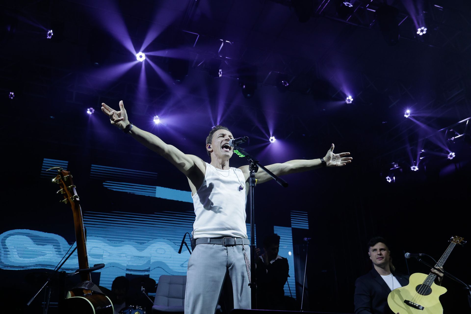 Um artista cantando com os braços erguidos no palco, iluminado por holofotes roxos, guitarrista visível.