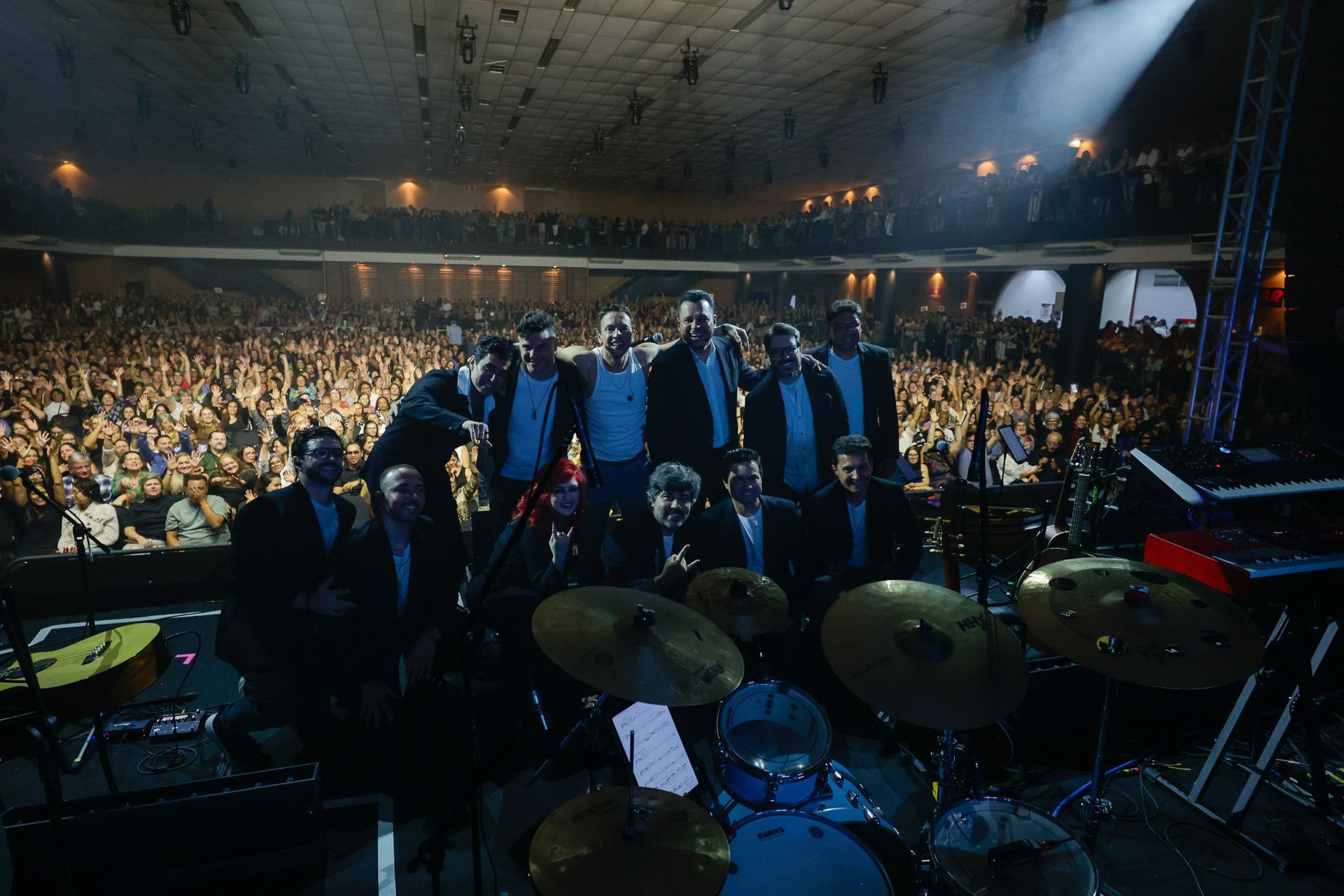Banda no palco com grande público em uma sala de concertos. Escuro, holofotes, instrumentos visíveis, pessoas aplaudindo.