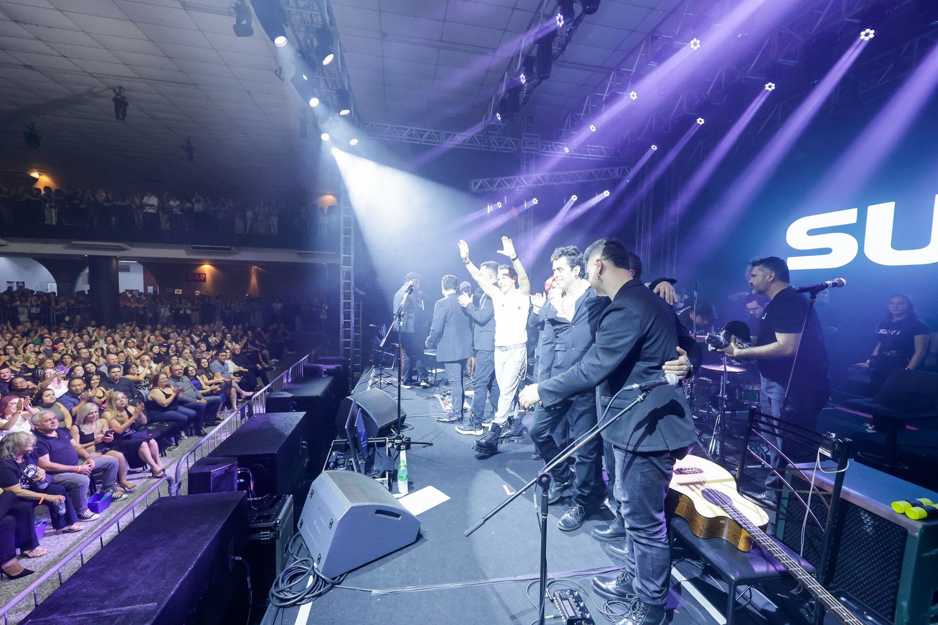 Banda no palco durante um concerto, apresentando-se para uma grande plateia em um local com luzes de palco brilhantes.