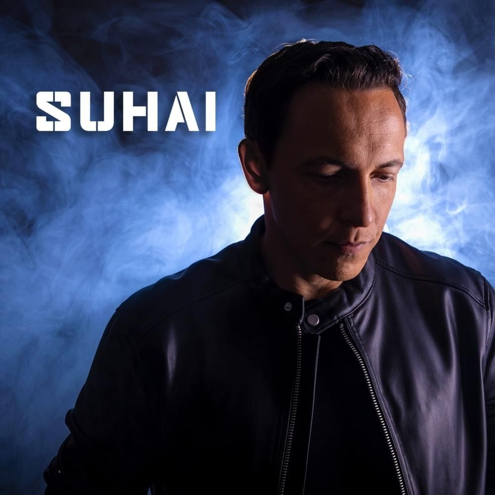 Homem chamado Suhai olhando para baixo, vestindo uma jaqueta preta, contra um fundo azul e esfumaçado.