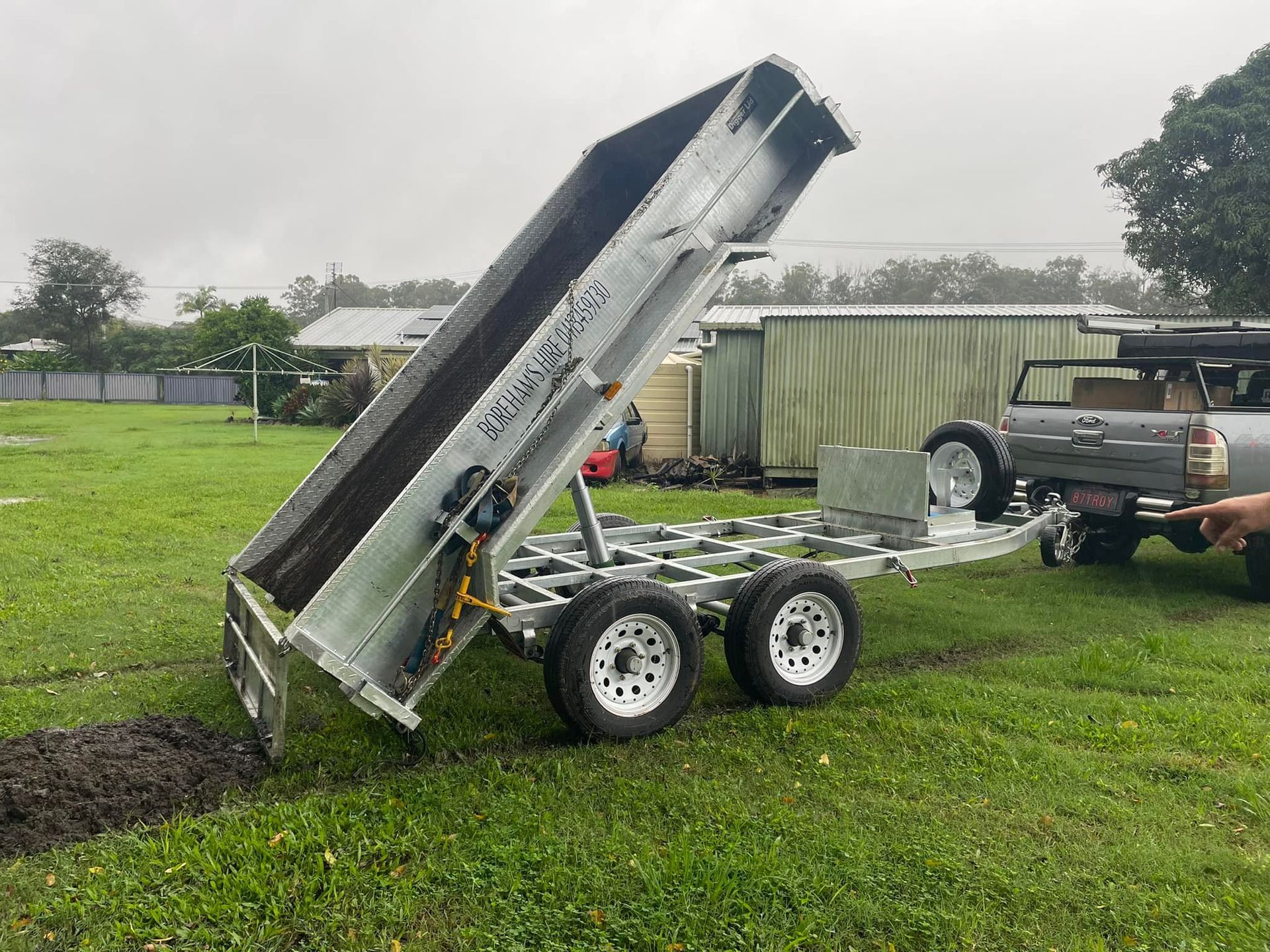 A Silver Tipper Truck With Its Bed Raised, Dumping a Dark Substance Onto Green Grass — Borehams Hire In Meridan Plains, QLD