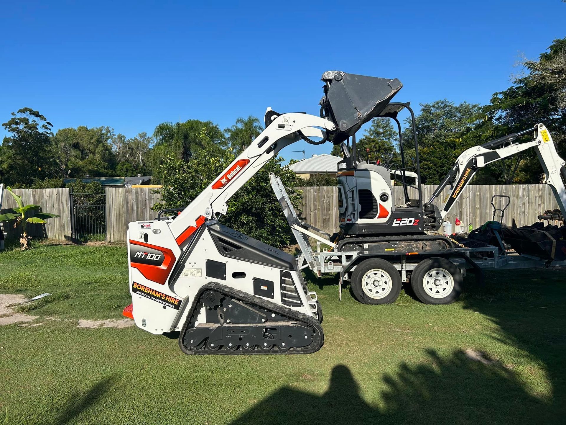 A White and Orange Track Skid Steer Next to an Excavator in a Grassy Yard — Borehams Hire In Meridan Plains, QLD