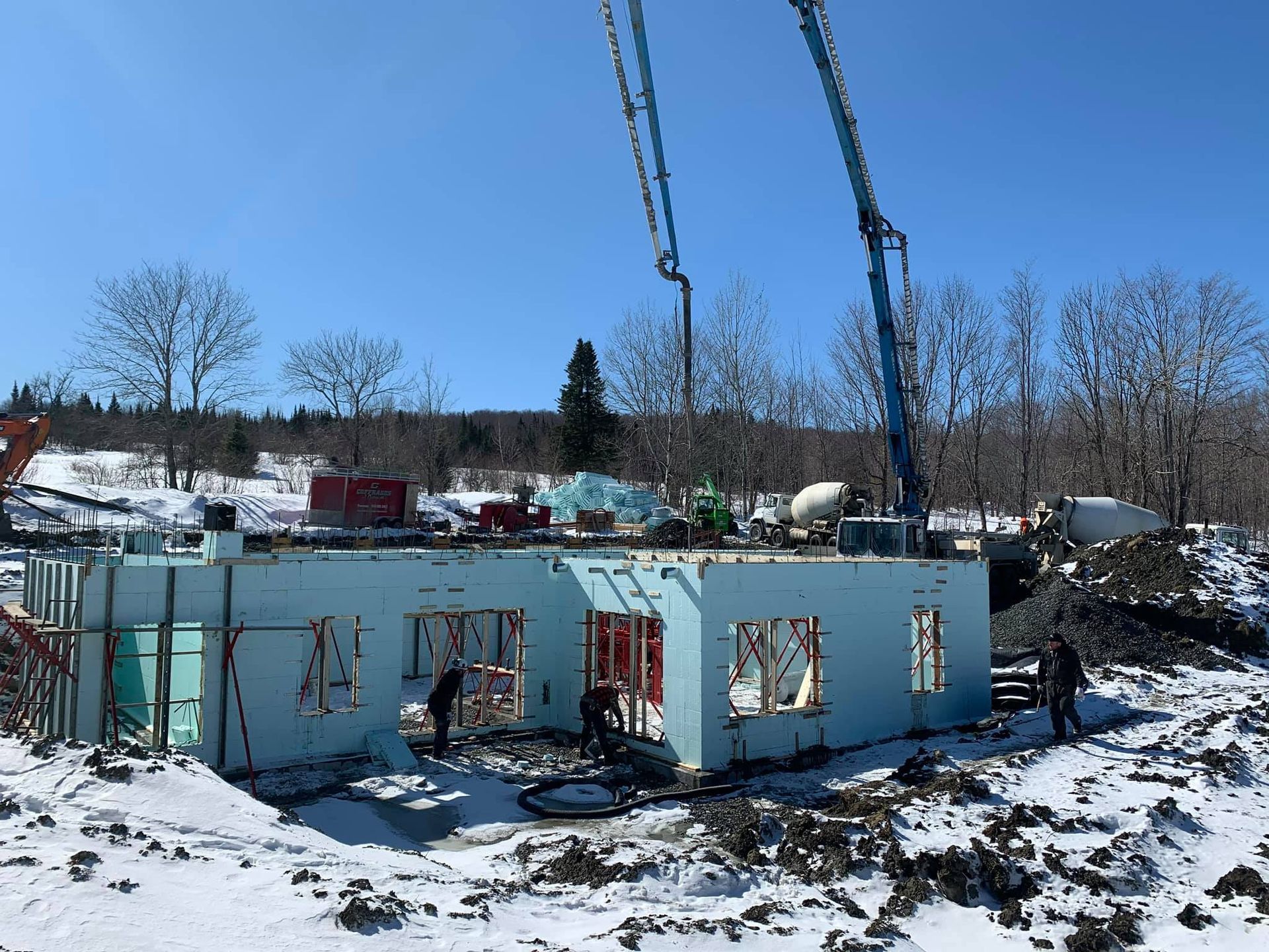 Chantier de construction avec murs en béton isolés bleu clair, neige et deux camions pompes à béton.