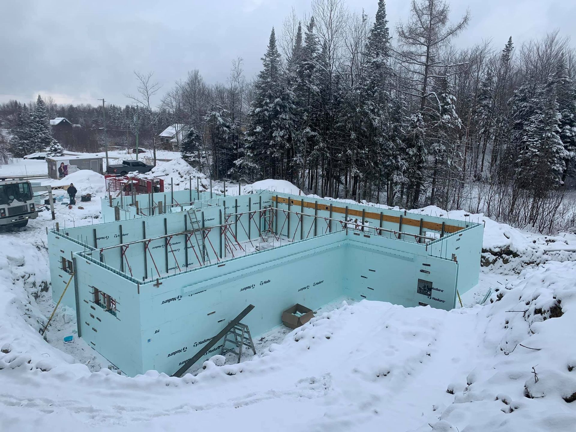 Chantier de construction avec murs en mousse bleue, barres d'armature et neige ; arbres en arrière-plan.