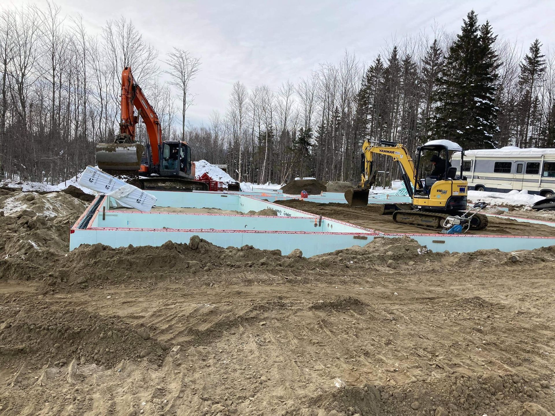 Chantier de construction avec excavatrices orange et jaunes, fondation en mousse bleue et sol enneigé.