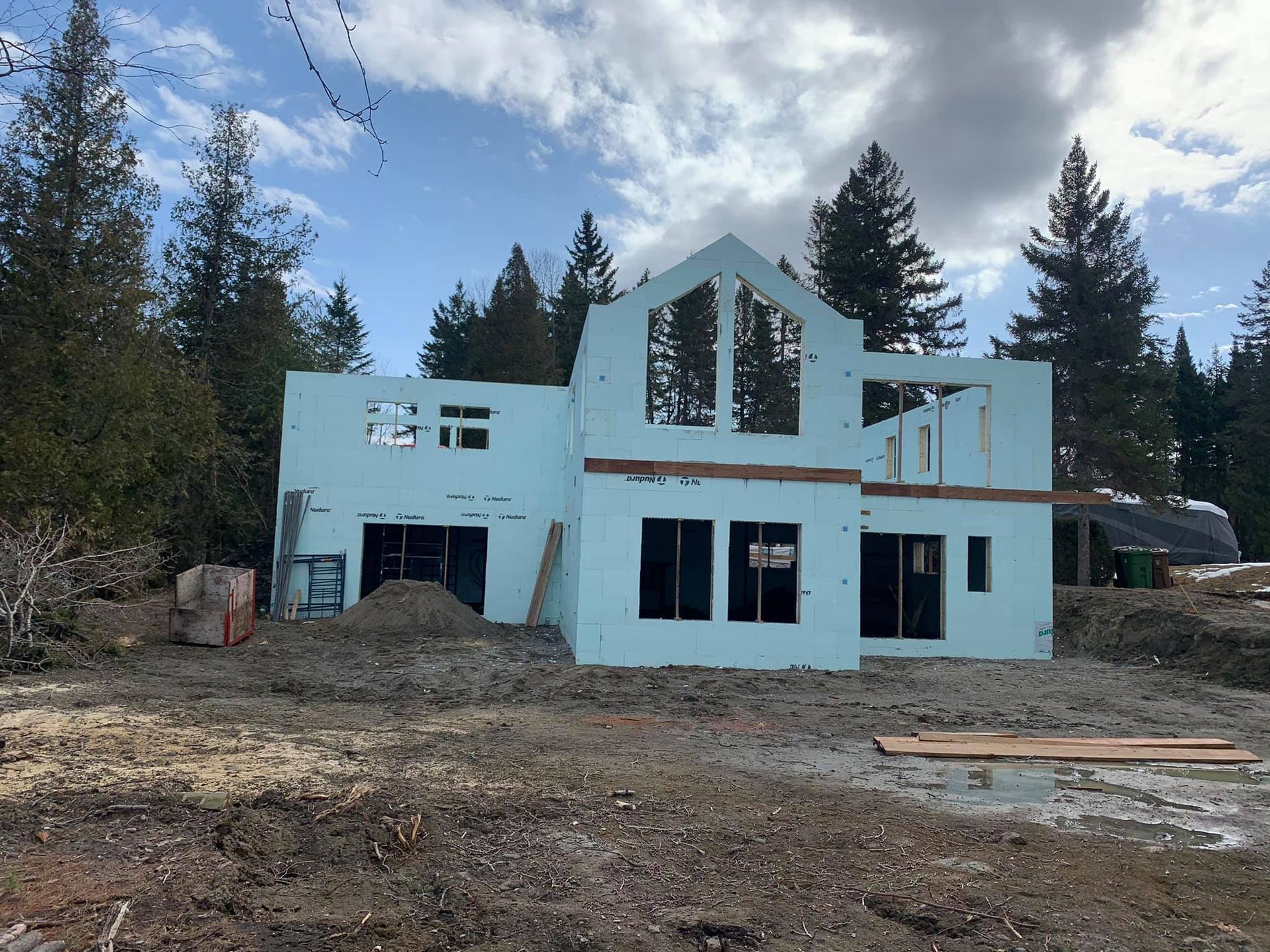 Une maison bleu clair partiellement construite sur un chantier entouré d'arbres. Ciel nuageux au-dessus.