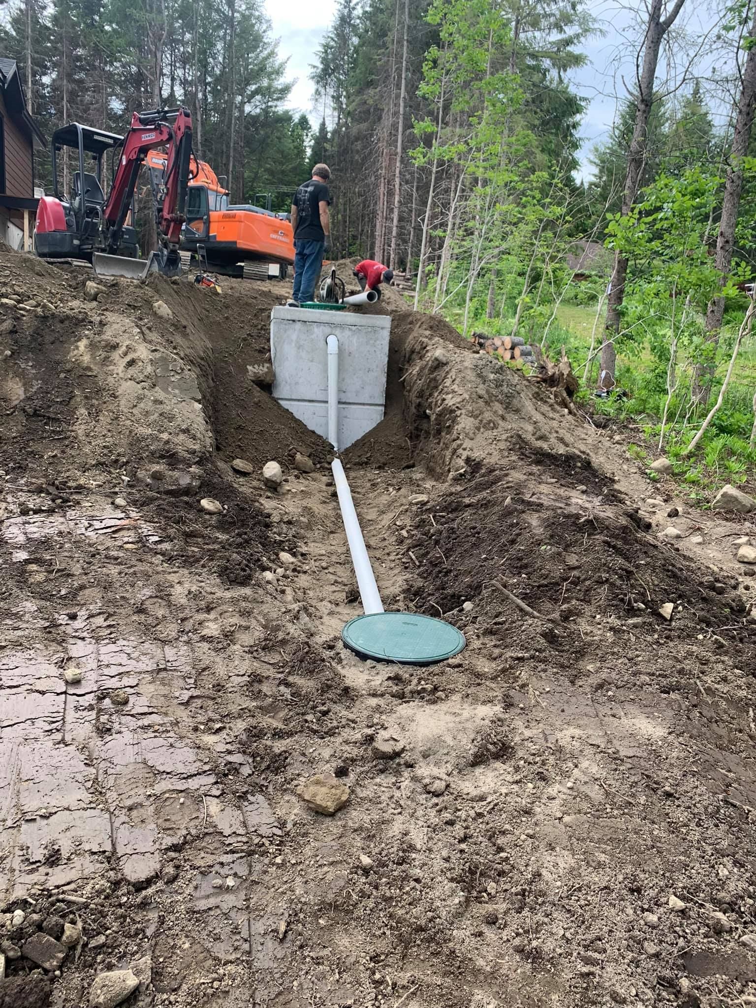 Chantier : Excavatrice, ouvriers et installation d'une fosse septique en béton dans une tranchée en terre, entourée d'arbres.