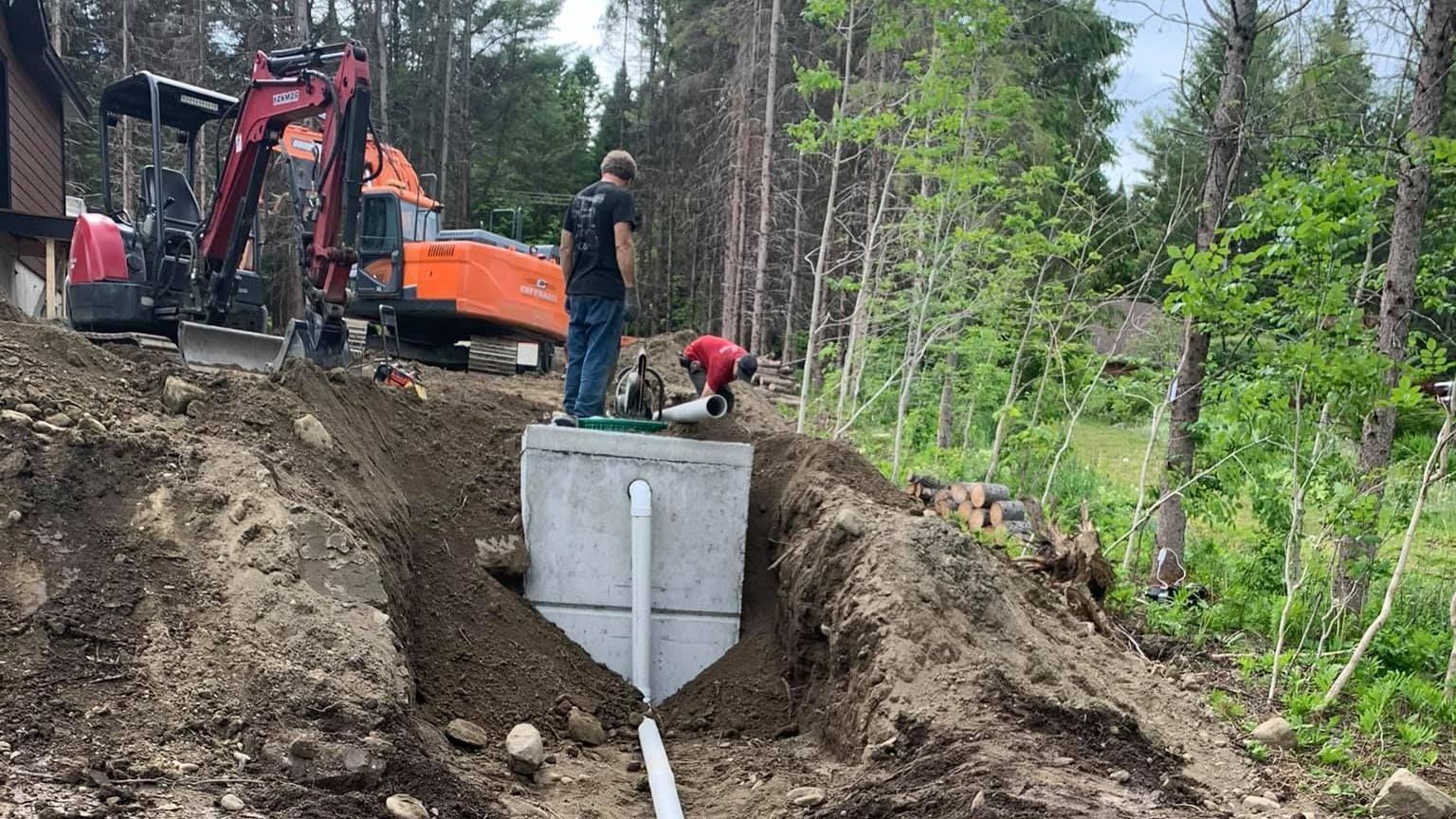 Des ouvriers installent une fosse septique dans une forêt, à l'aide d'une excavatrice, et raccordent des tuyaux.