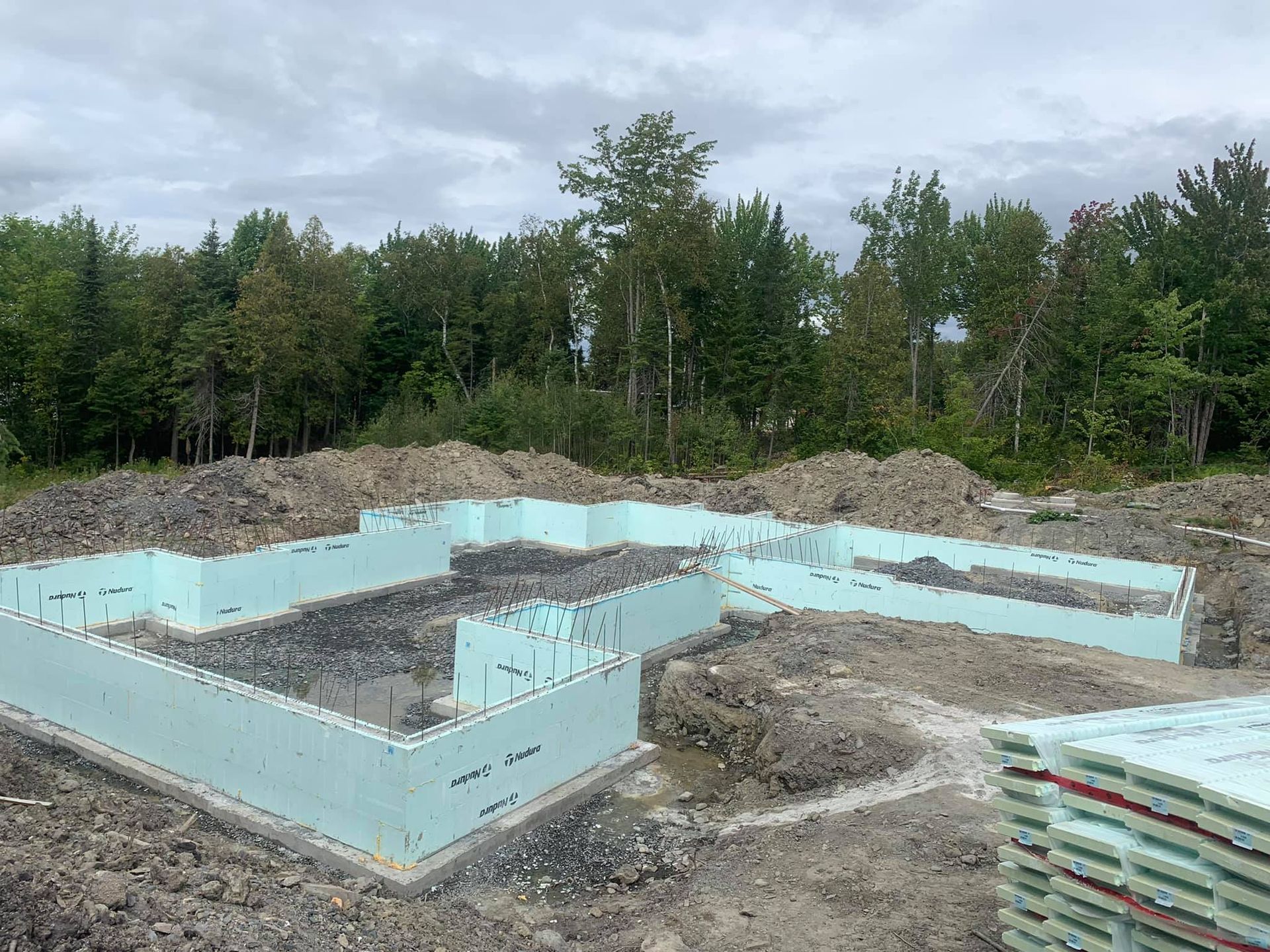 Murs de fondation en mousse bleue sur un chantier, entourés de tas de terre. Arbres verts en arrière-plan.