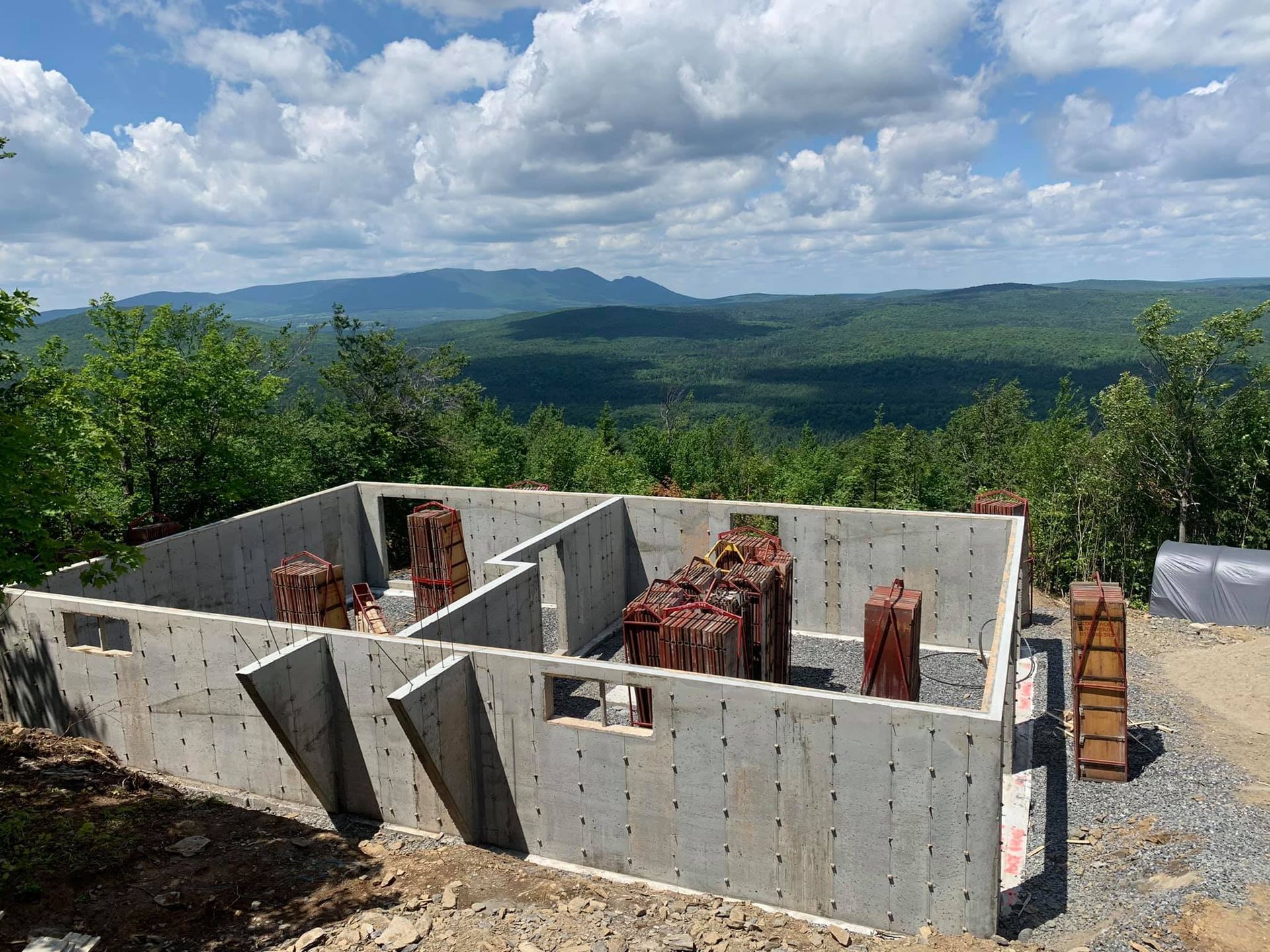 Fondation en béton avec barres d'armature apparentes, surplombant une chaîne de montagnes verdoyante sous un ciel nuageux.