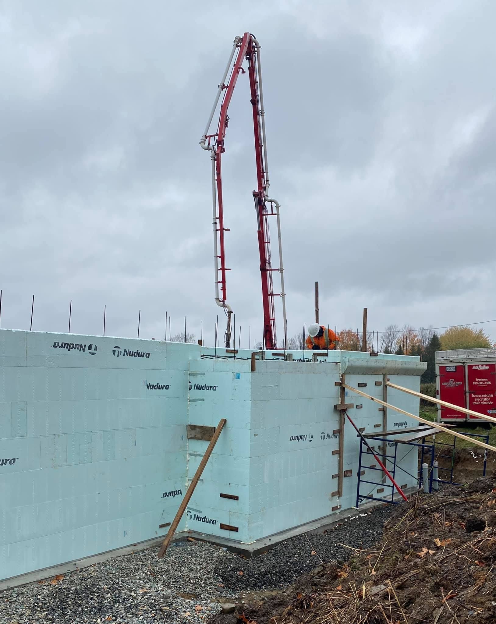 Chantier de construction avec du béton pompé dans un mur de mousse bleue, grue en arrière-plan, ciel couvert.