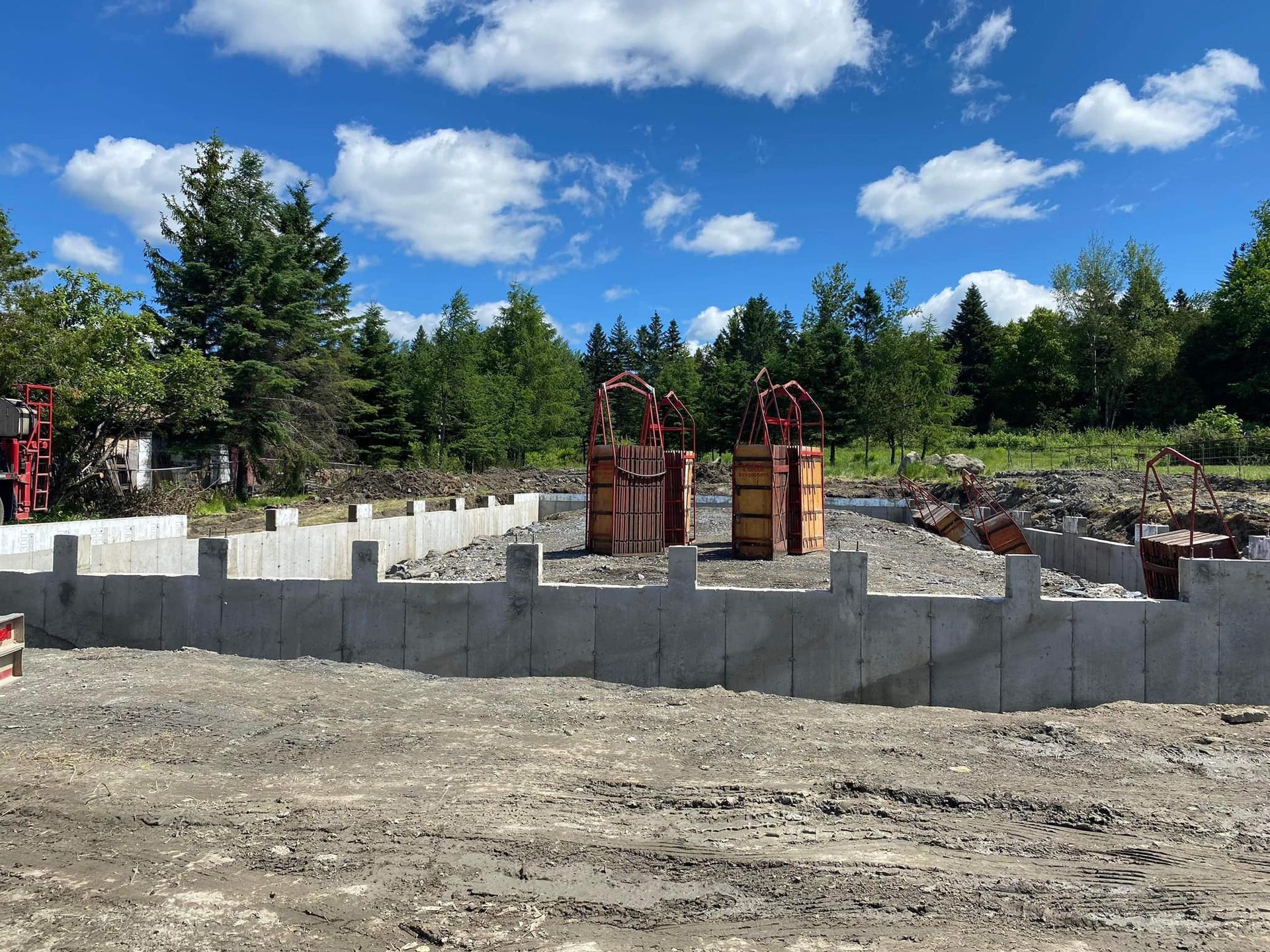 Chantier : fondation en béton, terre, arbres, ciel bleu avec nuages.