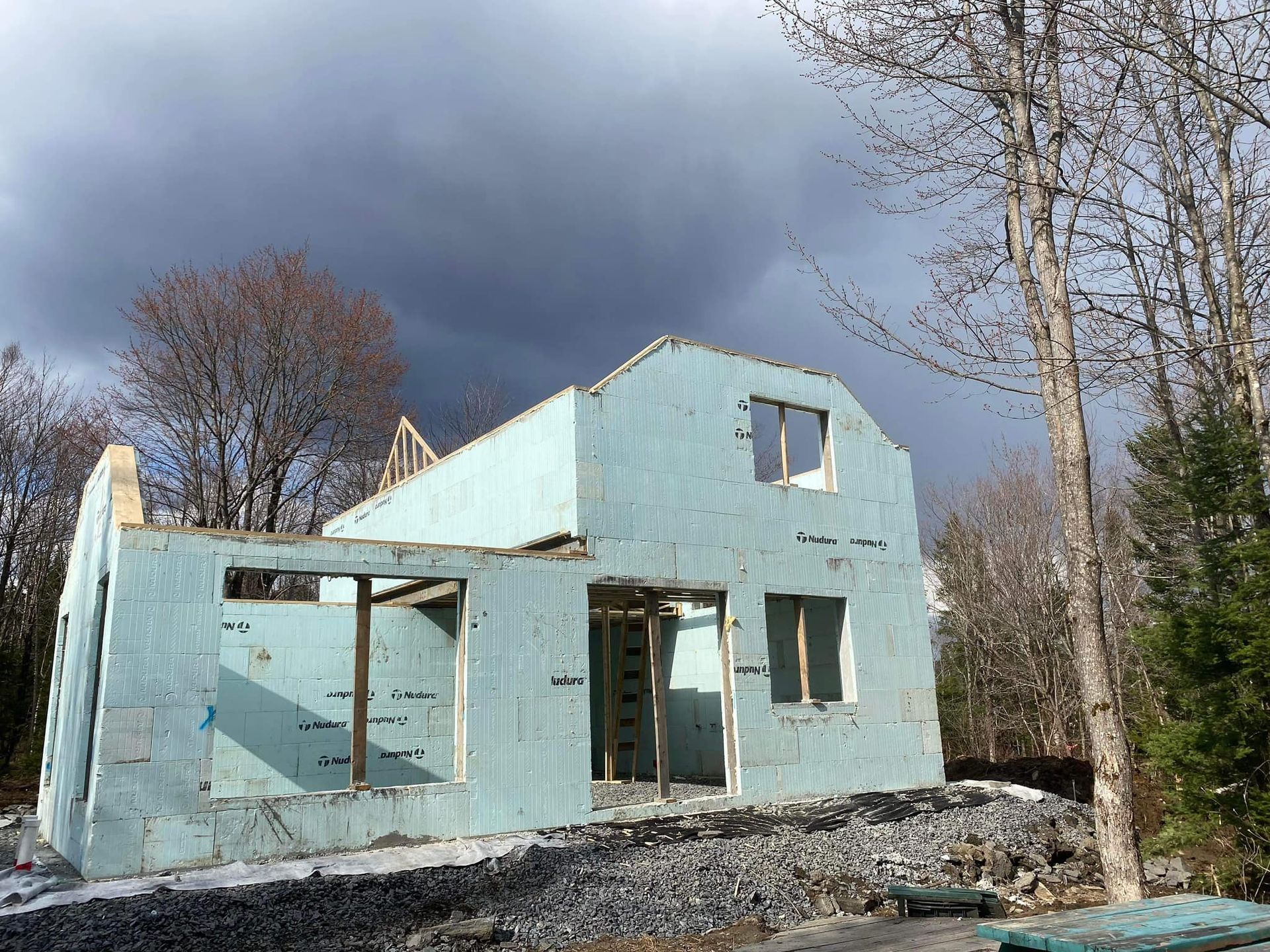Maison en construction avec des murs isolés bleus, se détachant sur un ciel nuageux et des arbres.