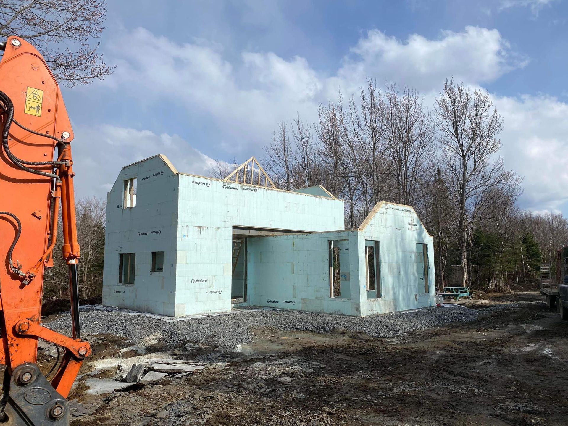 Chantier de construction avec un cadre de bâtiment en mousse bleu sarcelle, des arbres et une excavatrice orange.