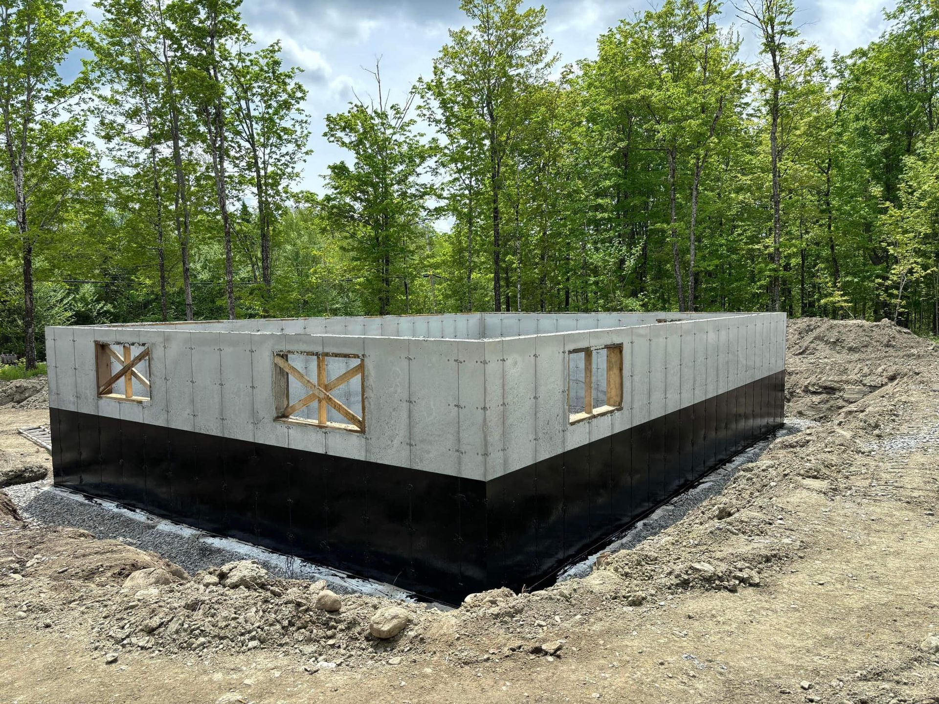 Fondation en béton d'un bâtiment en construction, entourée de terre et de gravier ; arbres en arrière-plan.