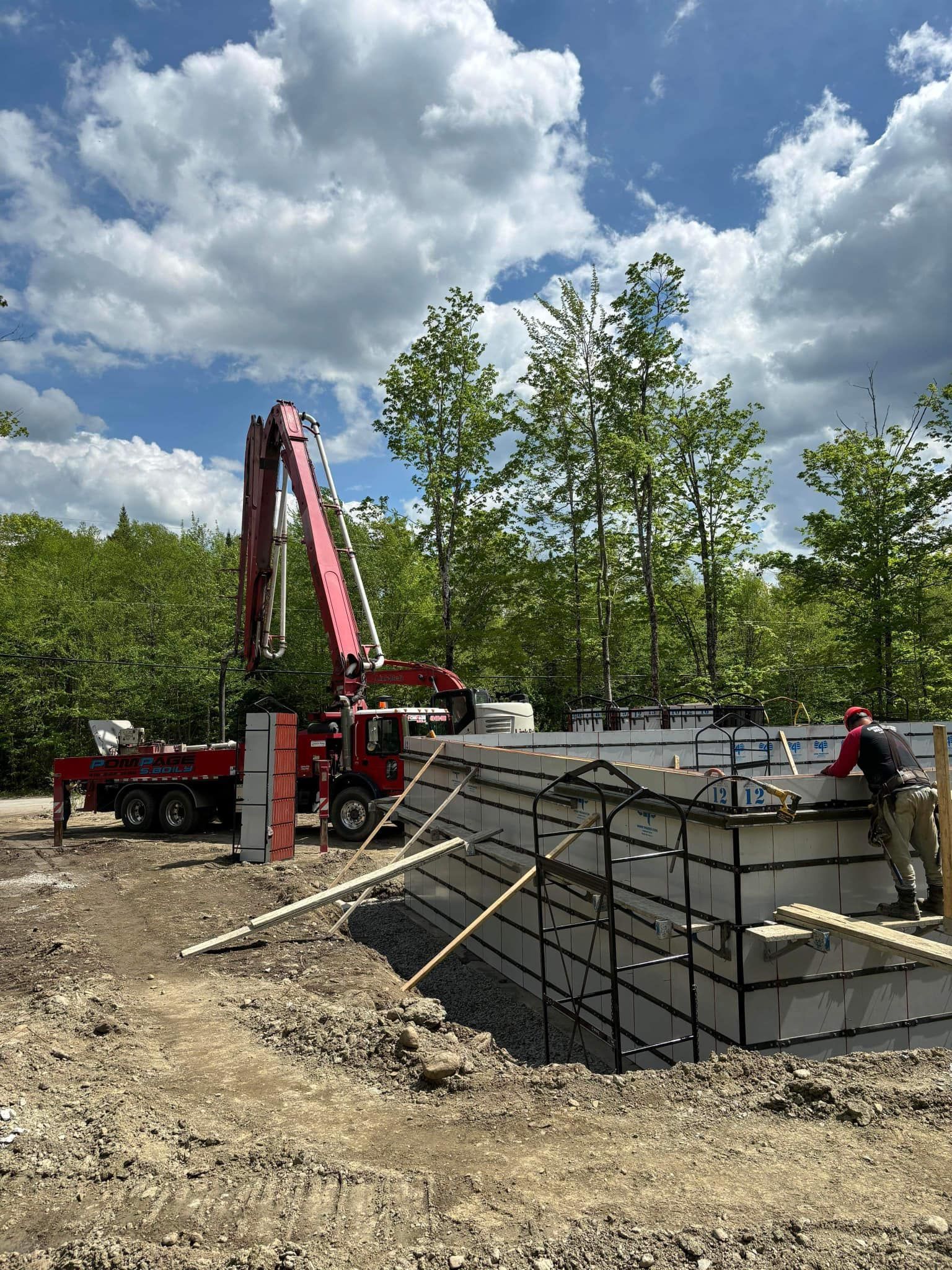 Chantier de construction : Camion pompe à béton coulant du béton dans des coffrages de fondation ; ouvrier en chemise rouge.