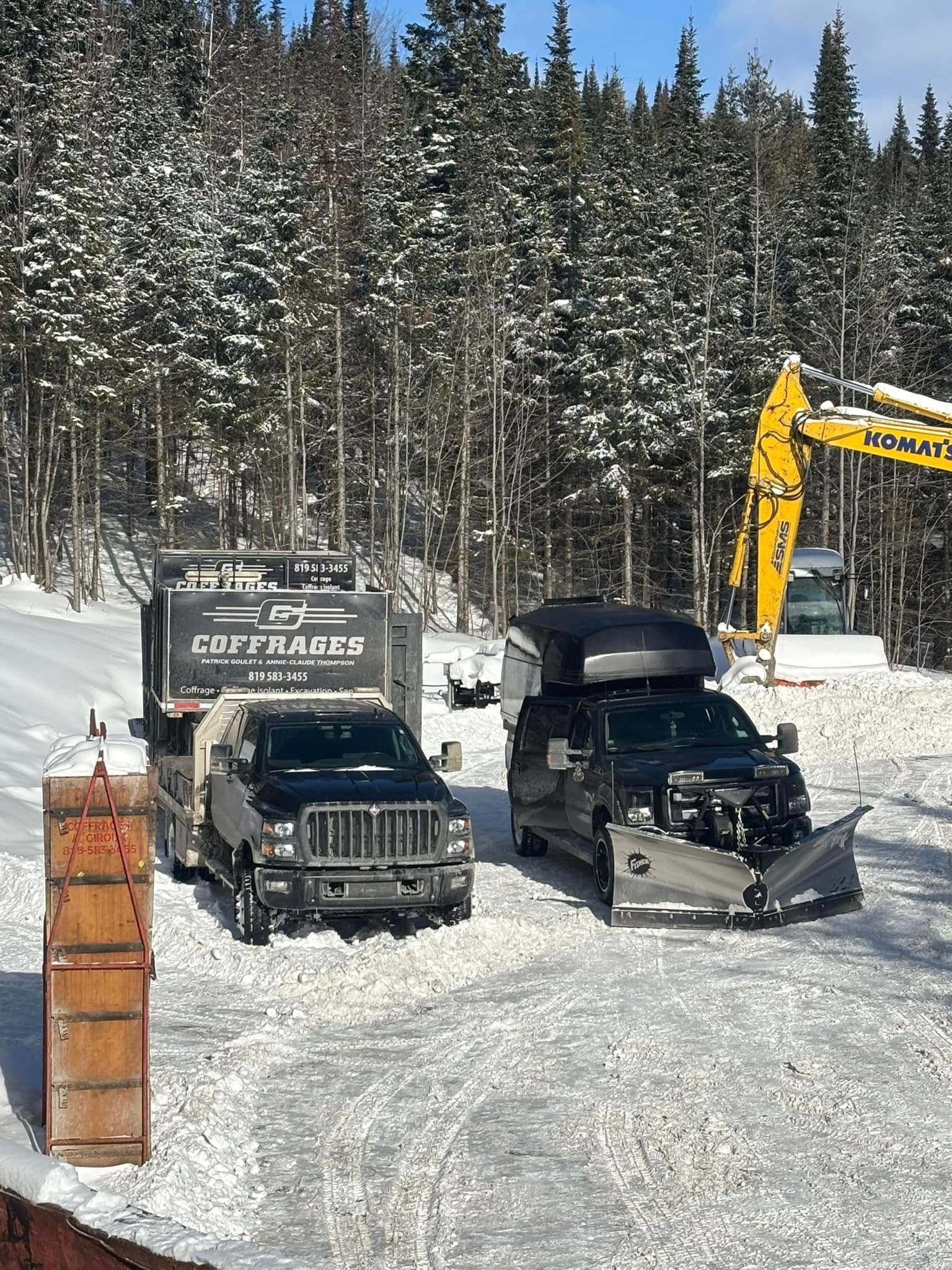 Deux camions noirs et une remorque dans une forêt enneigée, avec une excavatrice jaune en arrière-plan.
