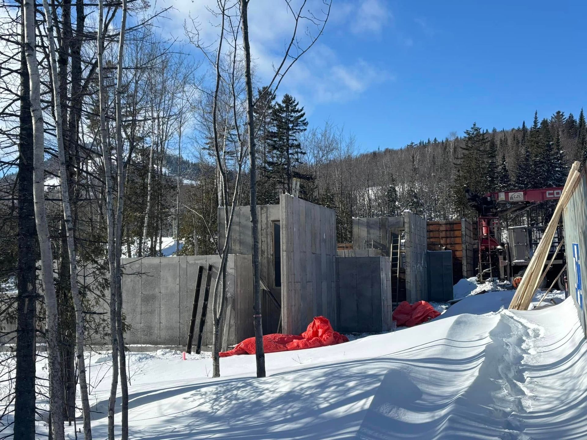 Chantier de construction dans un paysage enneigé : murs en béton, débris orange, arbres, ciel bleu.
