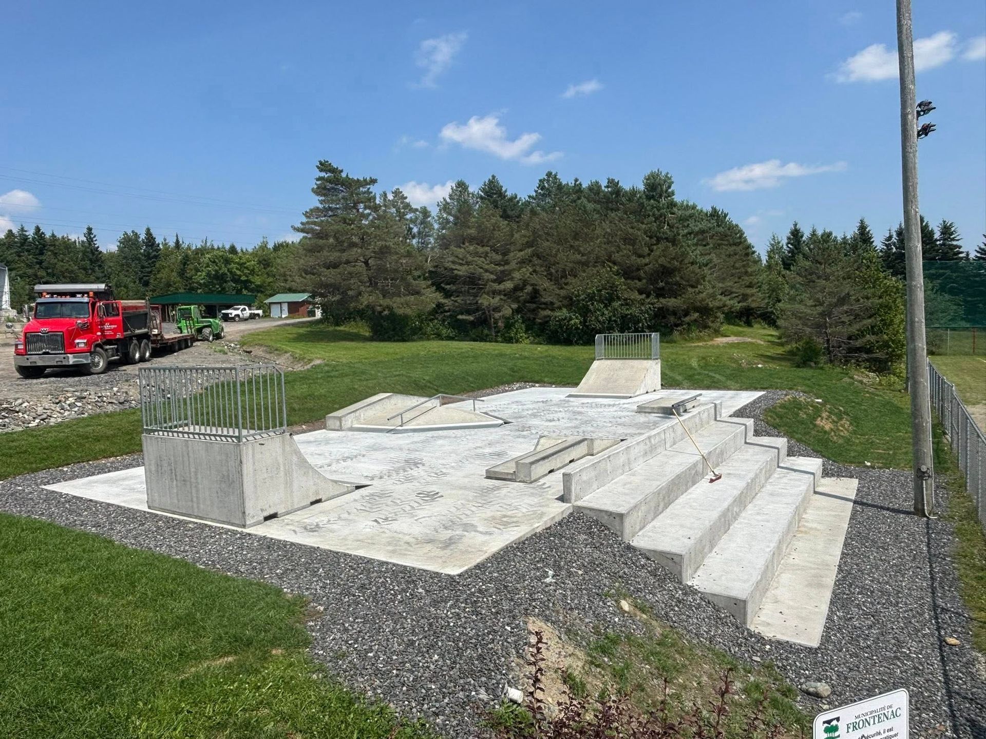 Skatepark en béton en construction avec rampes et marches, posé sur de l'herbe verte sous un ciel bleu.