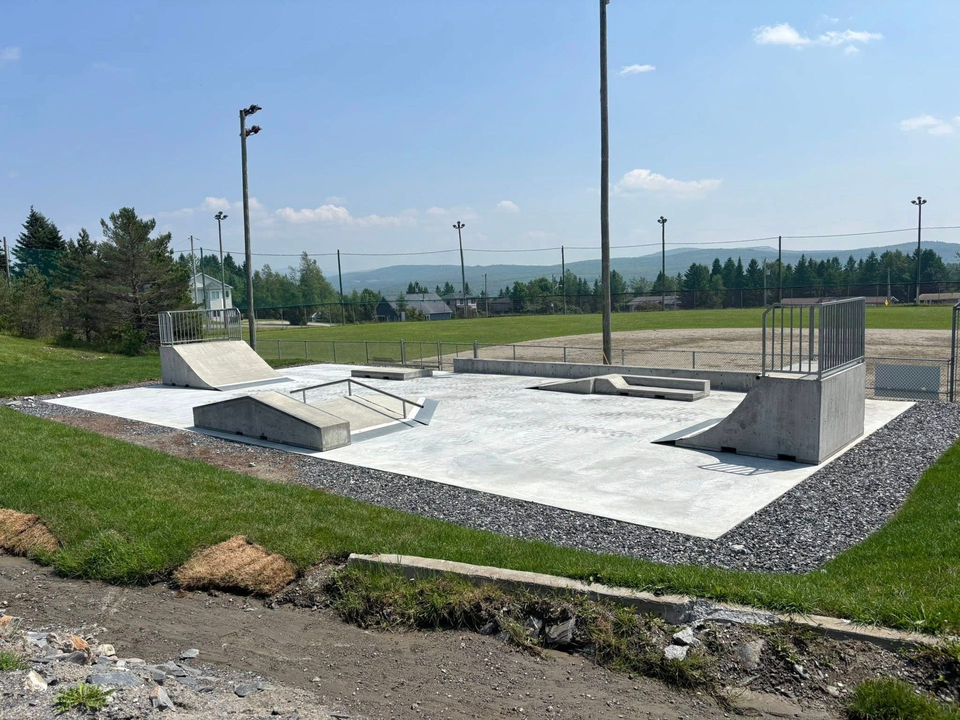 Un skatepark en béton avec rampes, rails et terrain plat. Le tout est entouré d'herbe verte.